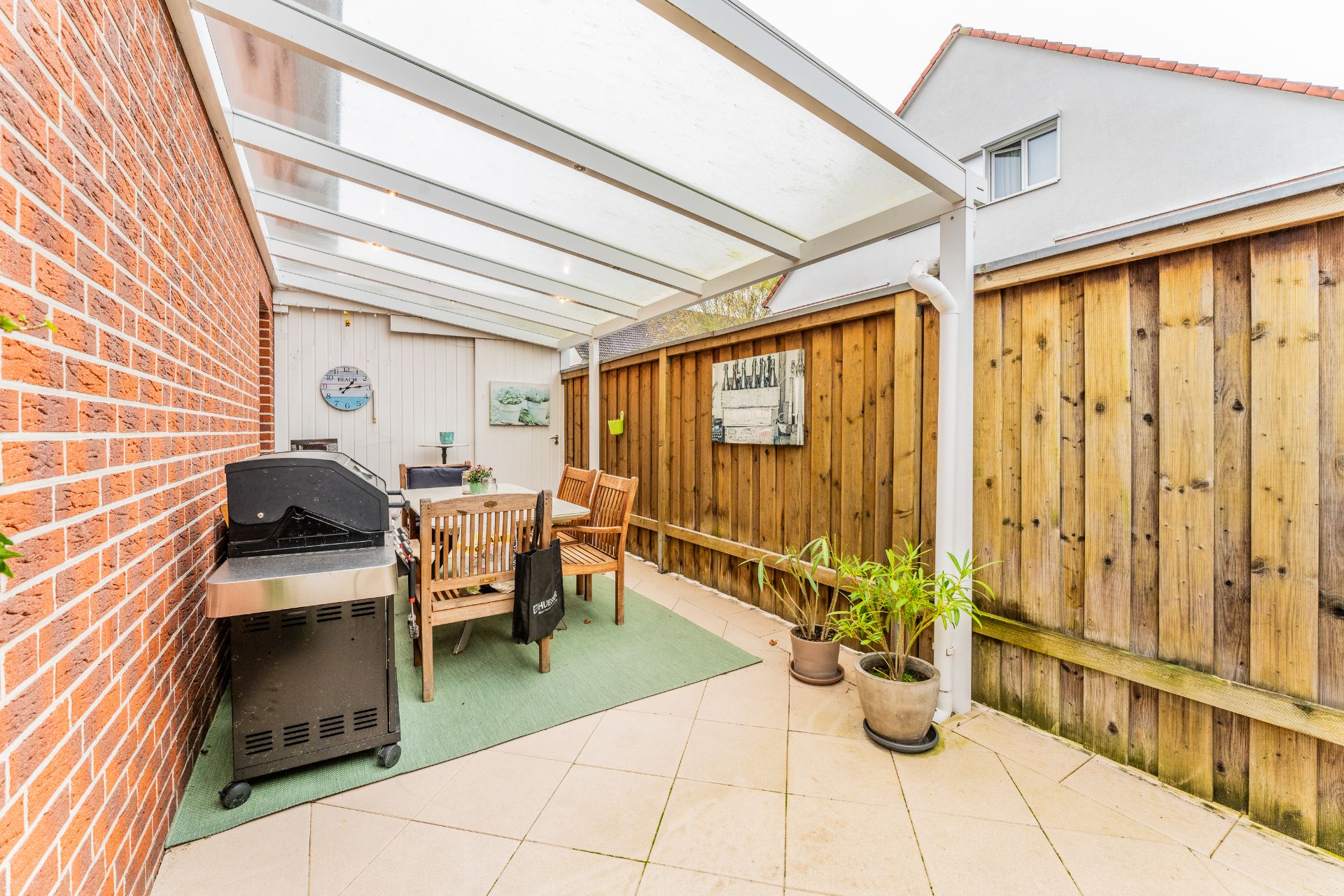 Überdachte Terrasse mit Grill, Holzmöbeln und Pflanzen neben einer Holzwand.