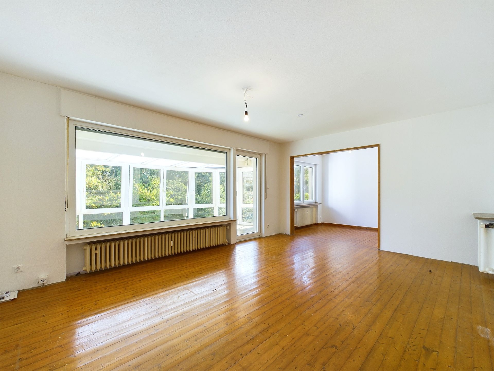 Helles Wohnzimmer mit Holzfußboden, großen Fenstern und Zugang zu einem weiteren Raum.