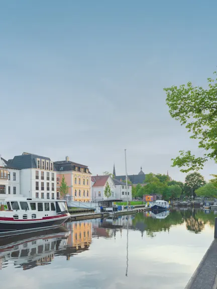 Mehrstöckige Wohnhäuser und Boote an einem ruhigen Kanal mit Bäumen im Hintergrund.