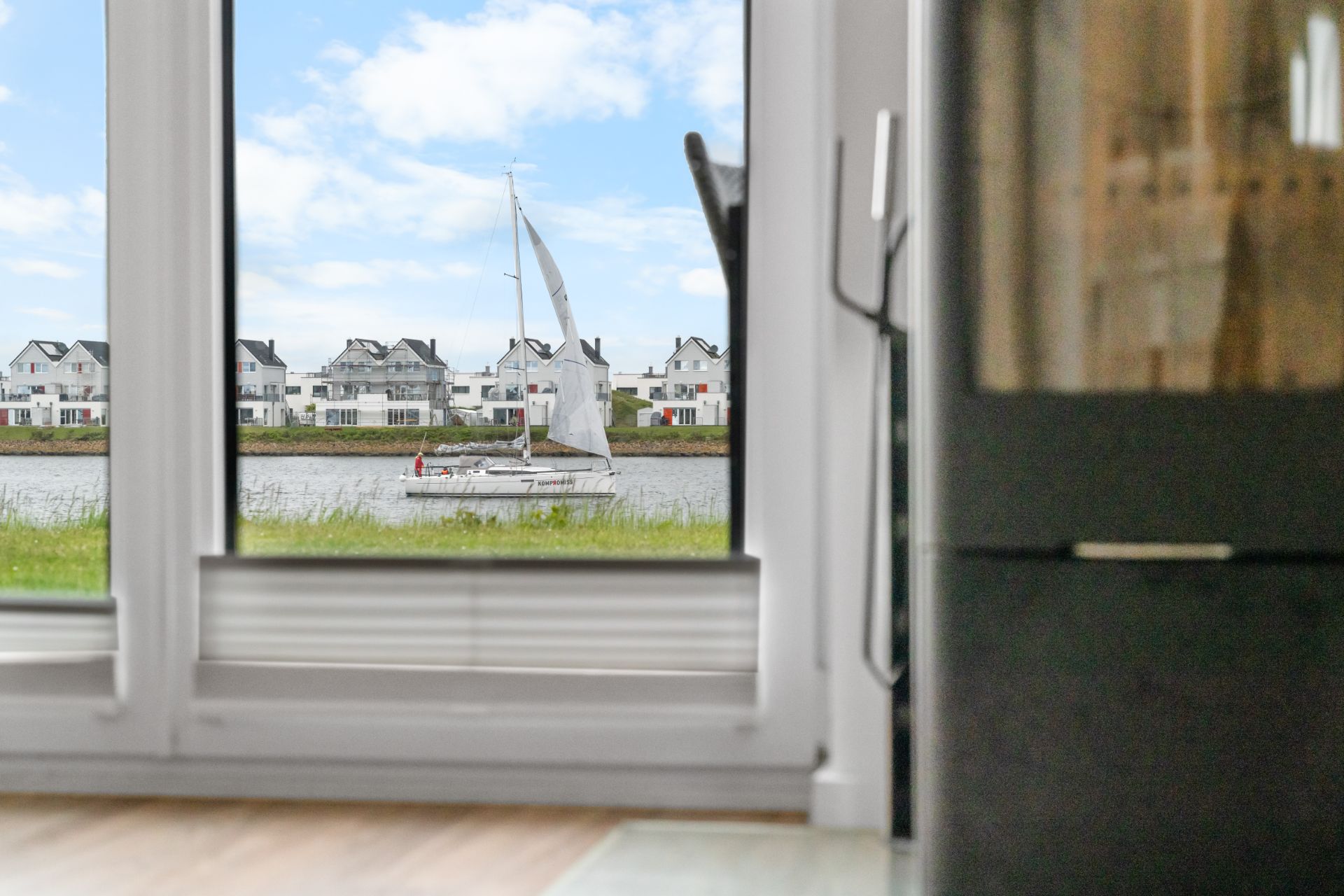 Blick durch große Fenster auf ein Segelboot vor Häusern am Wasser.