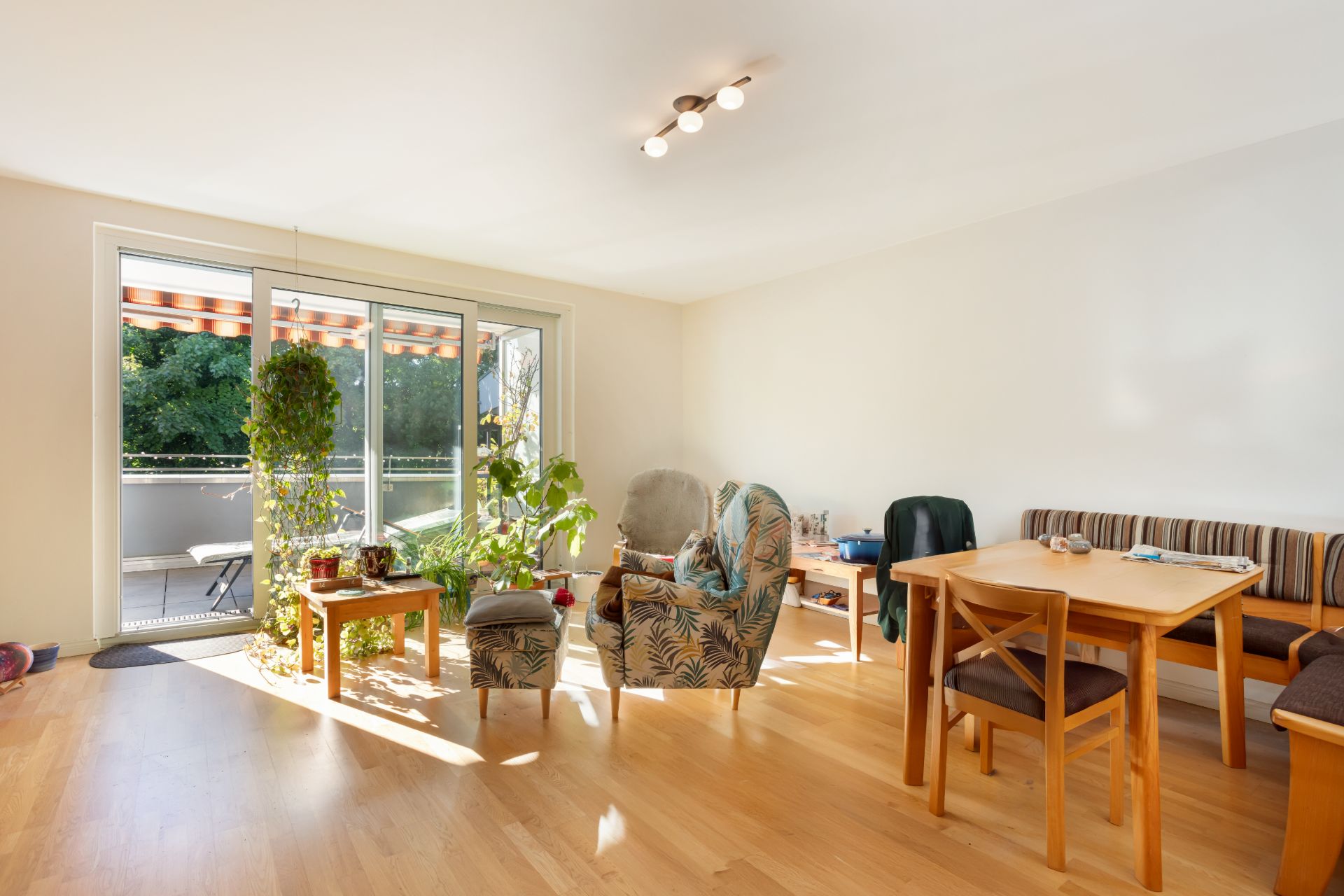 Helles Wohnzimmer mit Holzboden, Esstisch, Sesseln, Pflanzen und Zugang zum Balkon.