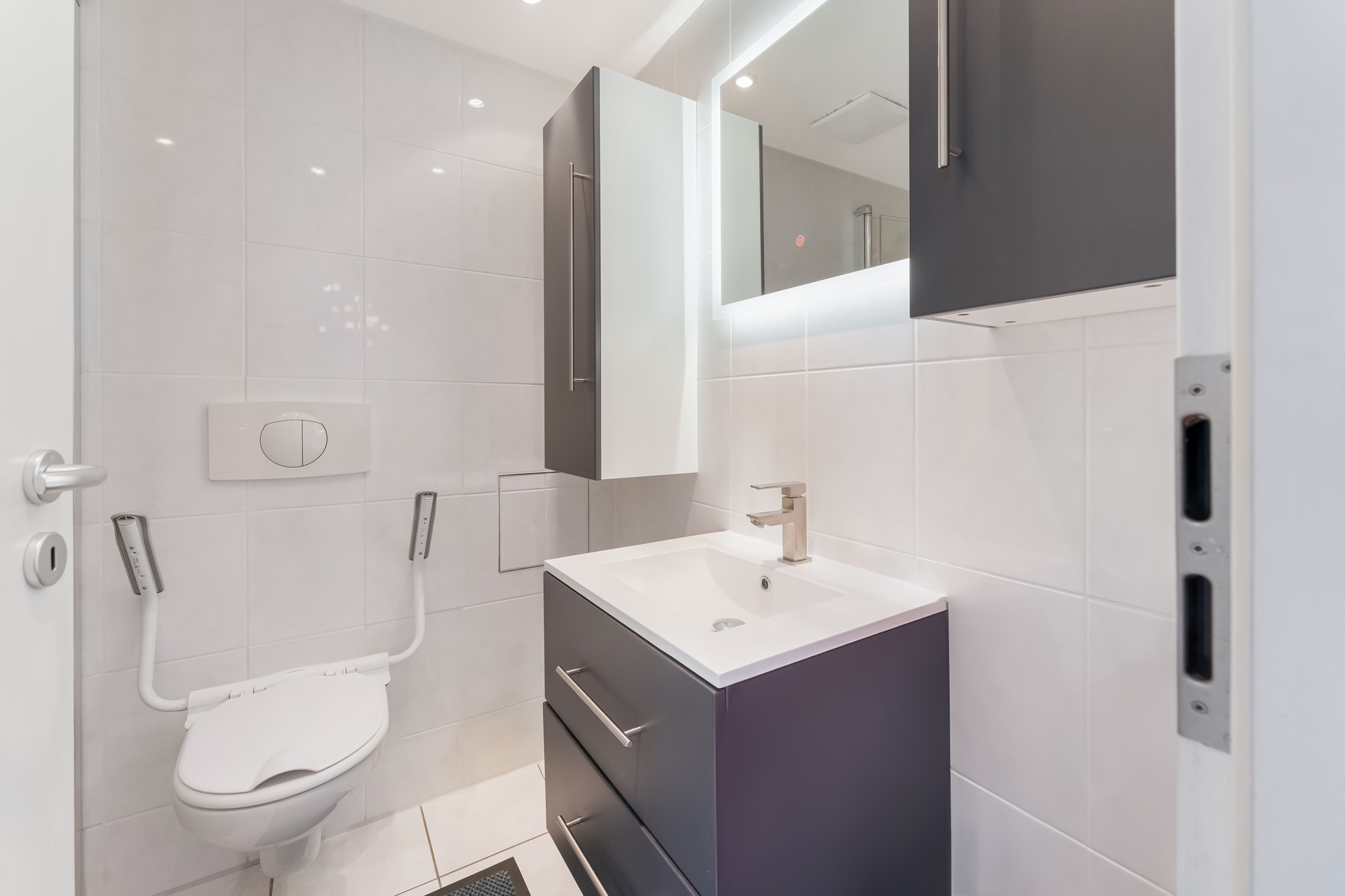 Modernes Badezimmer mit Wand-WC, Haltegriffen, Waschbecken und beleuchtetem Spiegel.