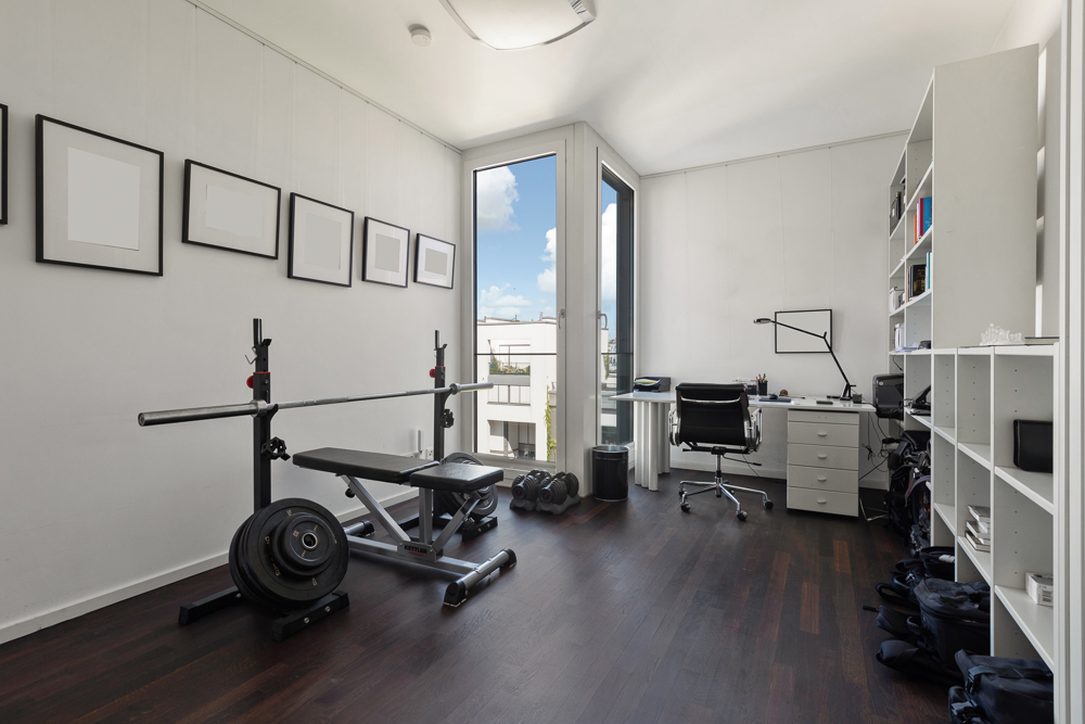 Helles Zimmer mit Hantelbank, Schreibtisch und weißen Regalen, großes Fenster mit Blick nach draußen.