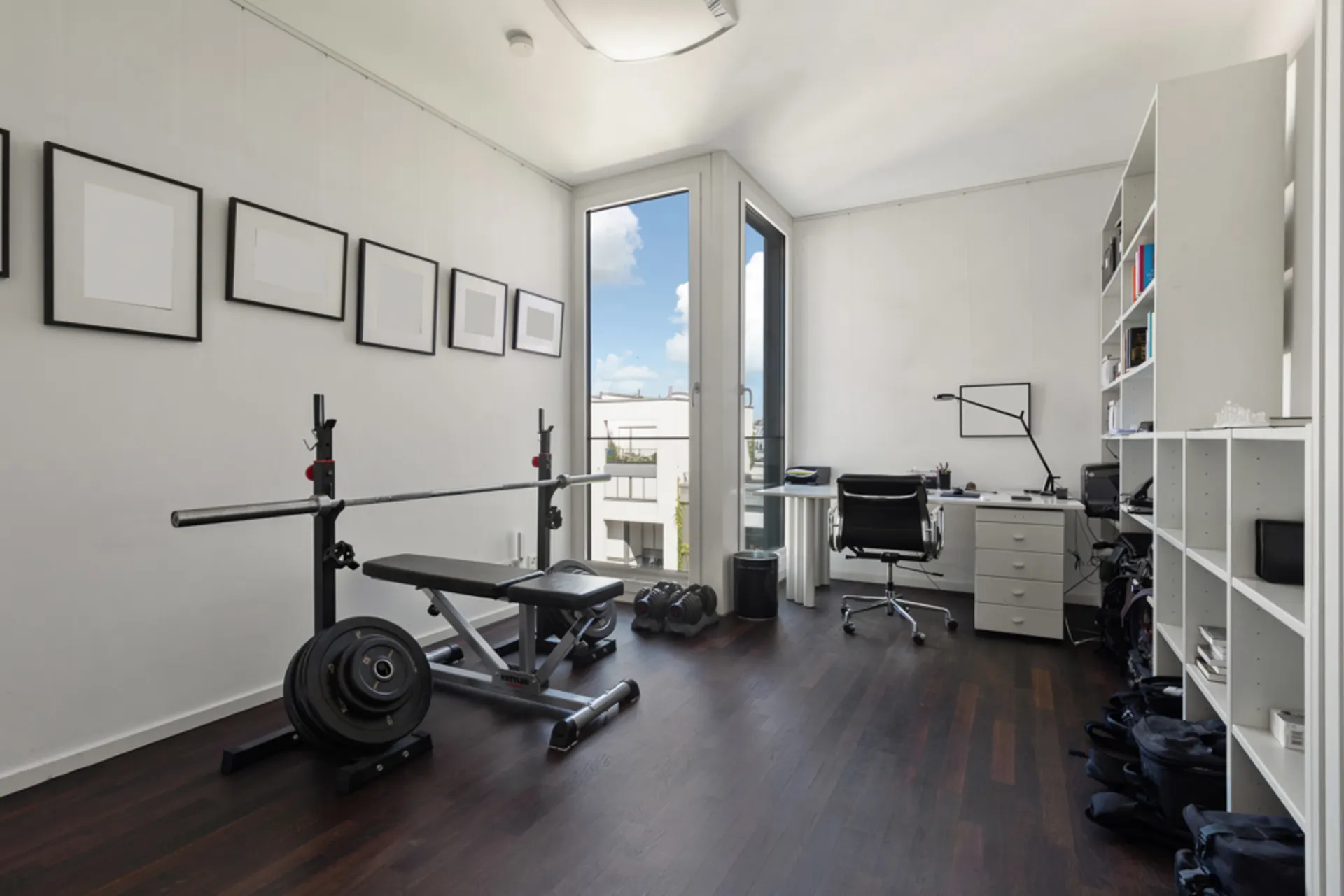 Helles Zimmer mit Hantelbank, Schreibtisch und weißen Regalen, großes Fenster mit Blick nach draußen.