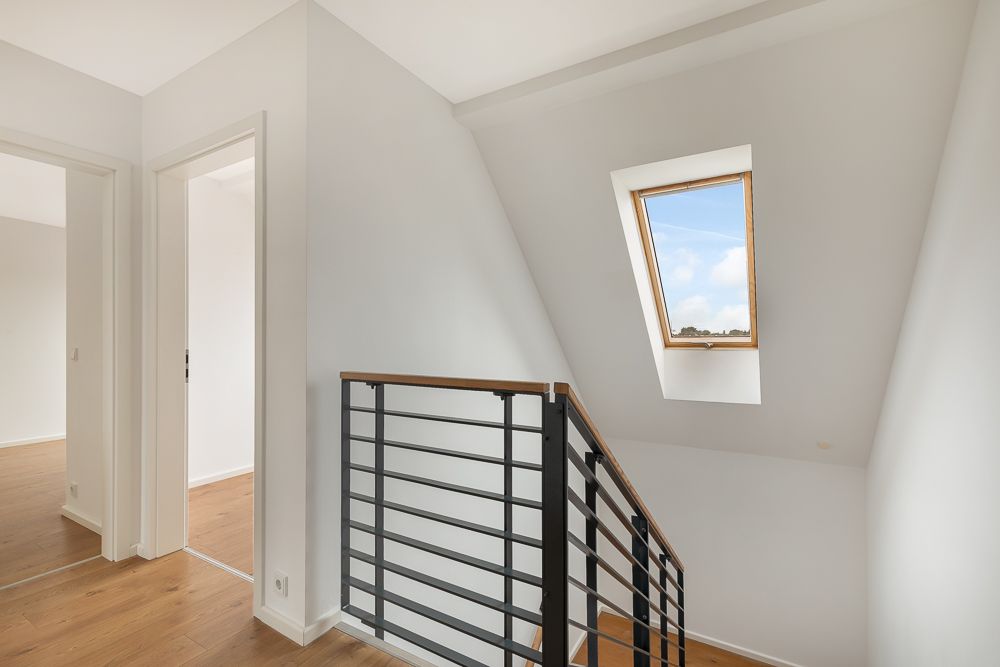 Heller Flur mit Treppengeländer, zwei Türen und Dachfenster mit Blick nach draußen.