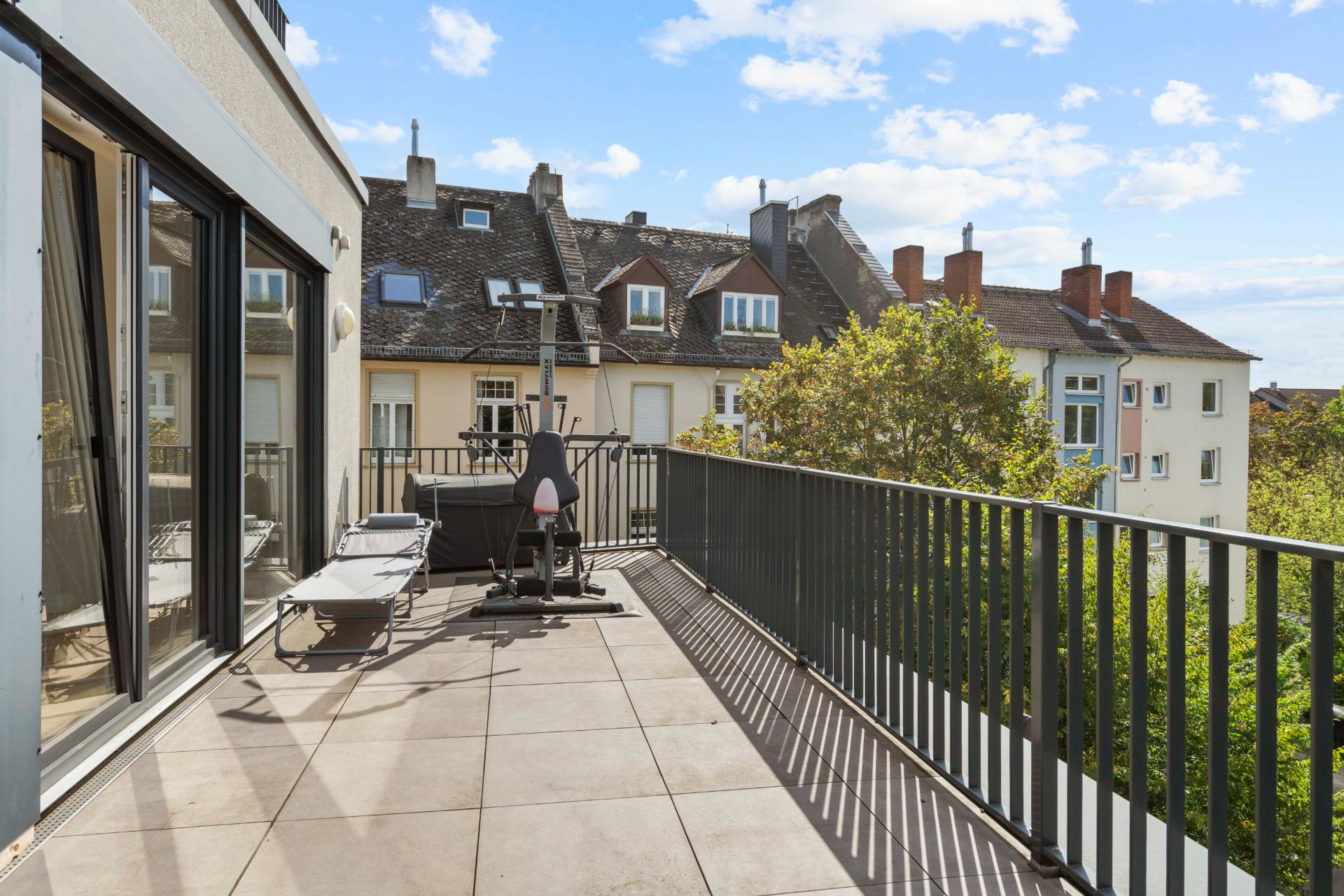 Großer Balkon mit Fitnessgerät, Liege und Blick auf Nachbarhäuser und Bäume.