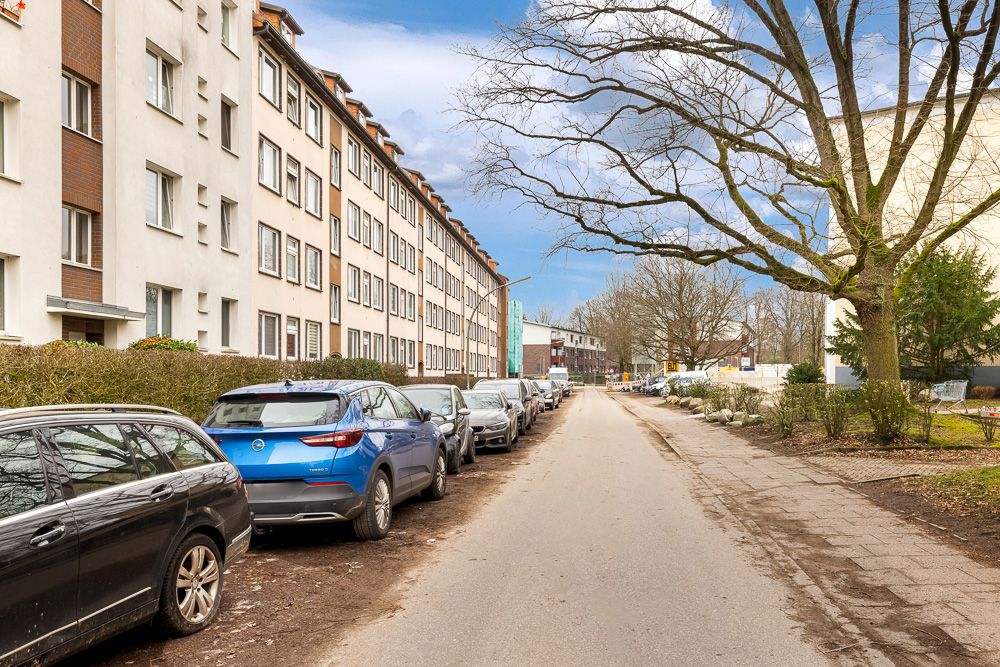 Wohnstraße mit parkenden Autos, mehrstöckigen Wohnhäusern und Bäumen am Straßenrand.