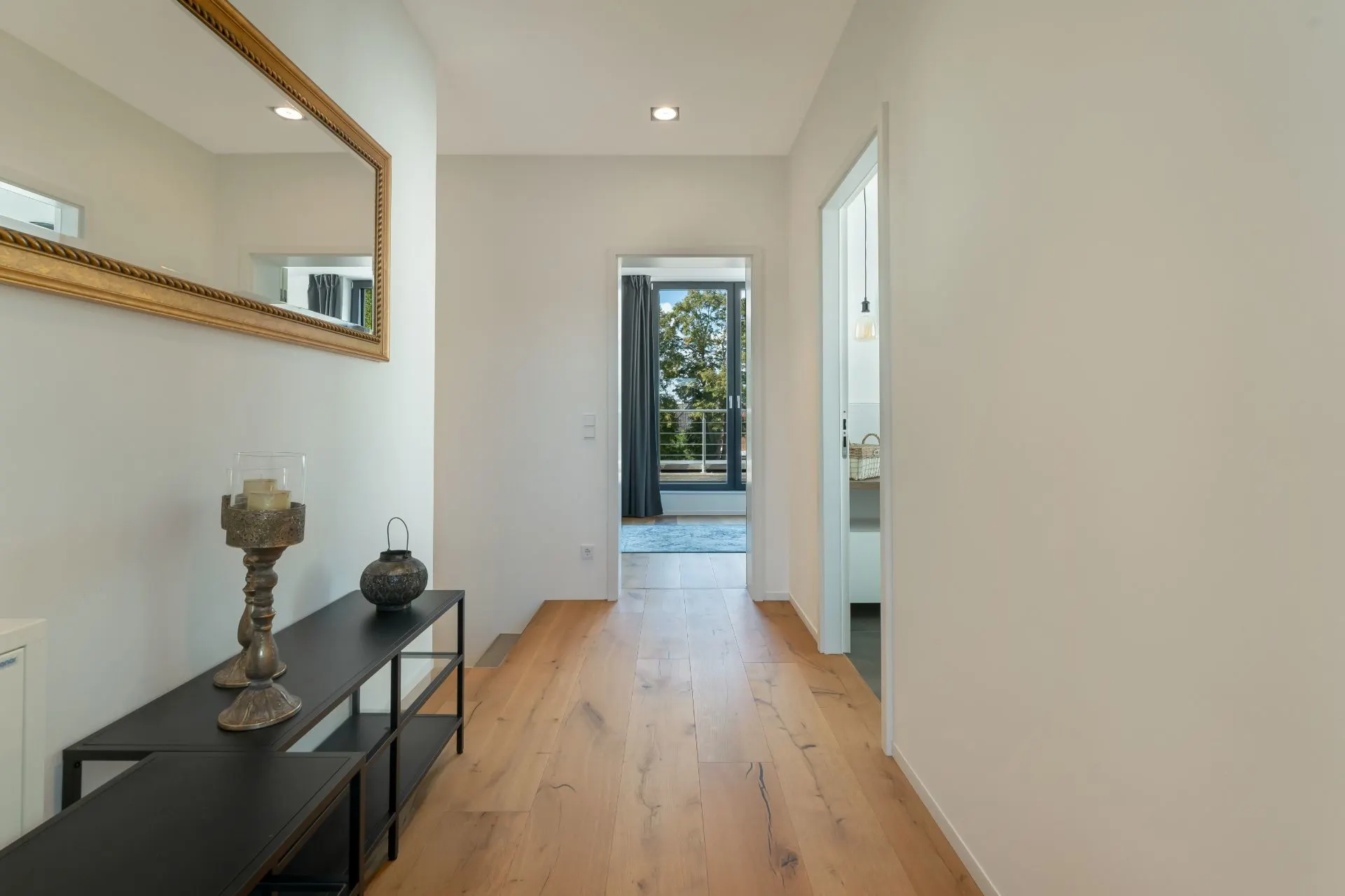 Heller Flur mit Holzfußboden, Spiegel und Dekoration, Blick ins nächste Zimmer mit Balkon.