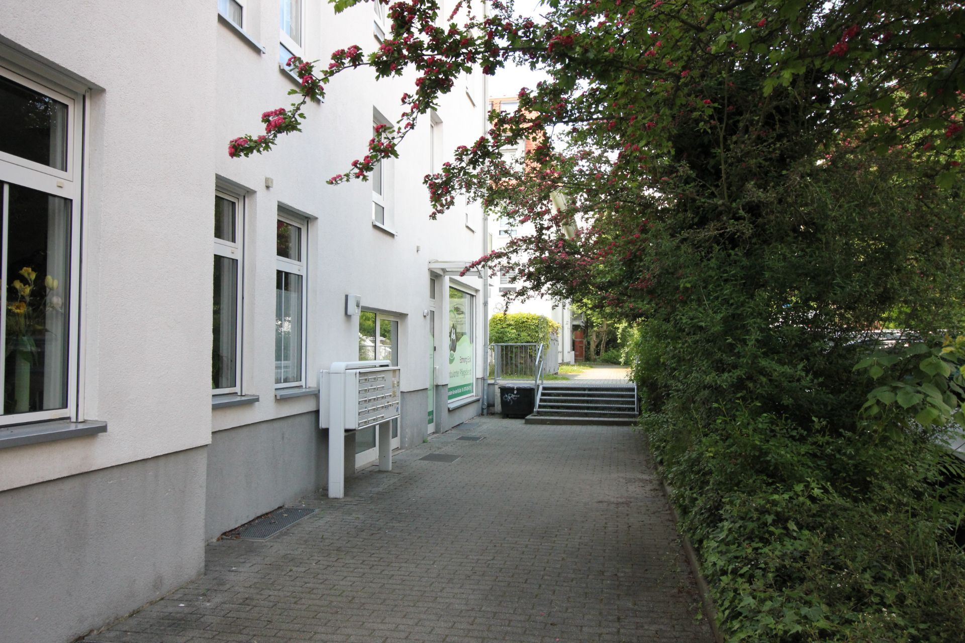Gepflasterter Weg neben Wohnhaus mit Briefkästen und angrenzenden Büschen.