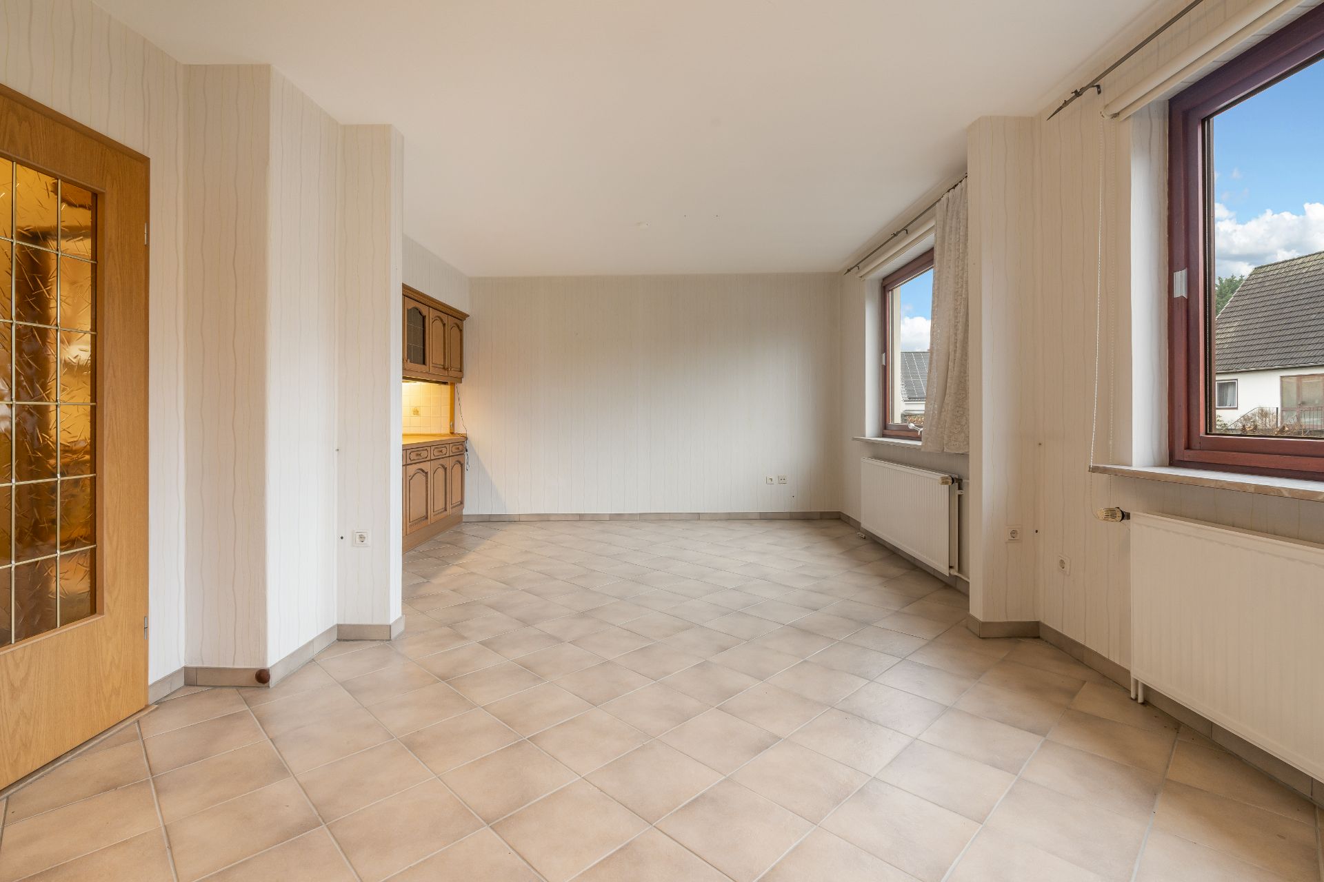 Leerer Raum mit gefliesten Boden, zwei Fenstern, Holztür und einer kleinen Einbauküche.