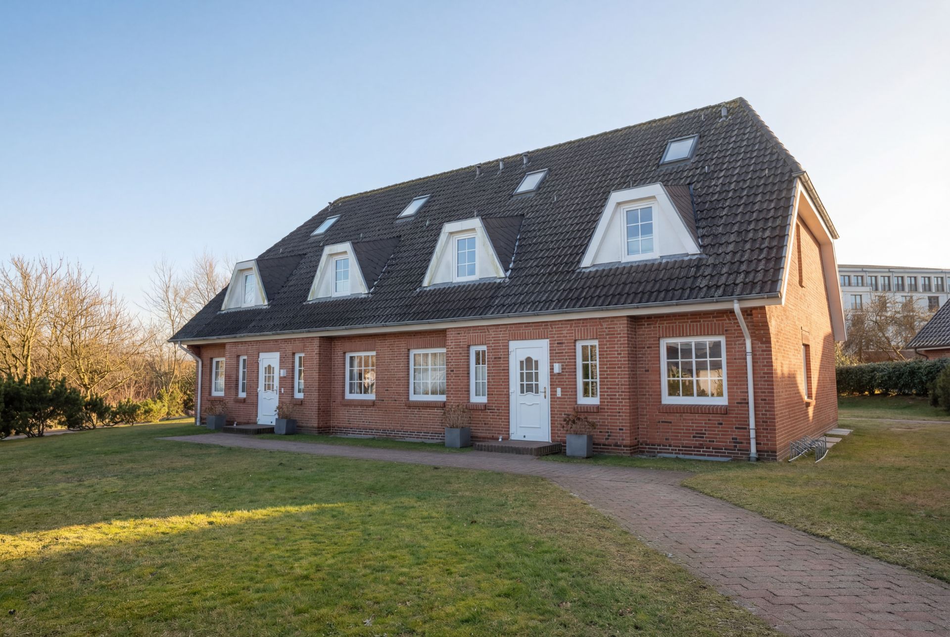 Backstein-Doppelhaus mit dunklem Dach und mehreren Dachfenstern, umgeben von Wiese.