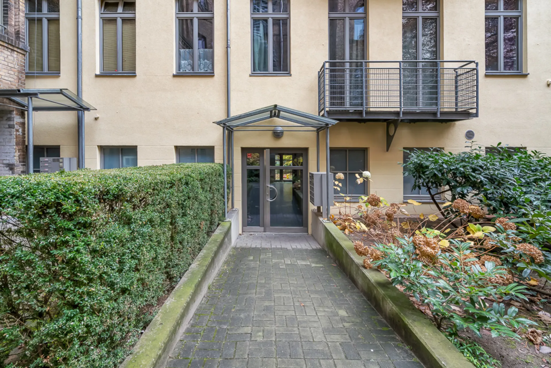 Eingang eines Wohngebäudes mit Glasdach, Hecken und Blumenbeeten seitlich des Weges.