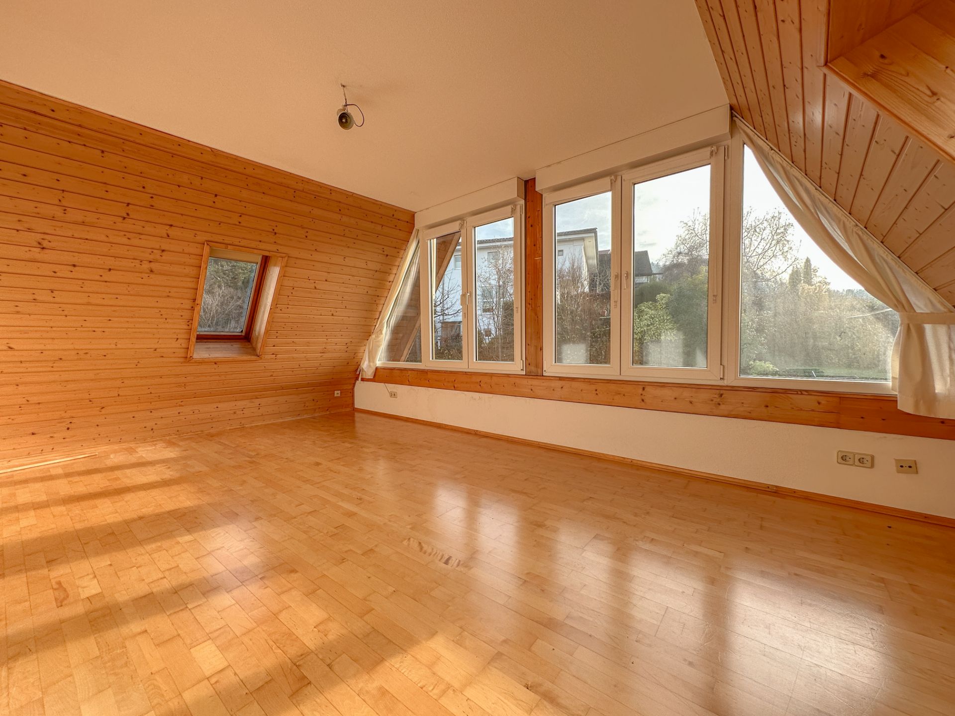 Leeres Zimmer mit Holzfußboden, holzverkleideten Wänden und großen Fenstern mit Blick nach draußen.
