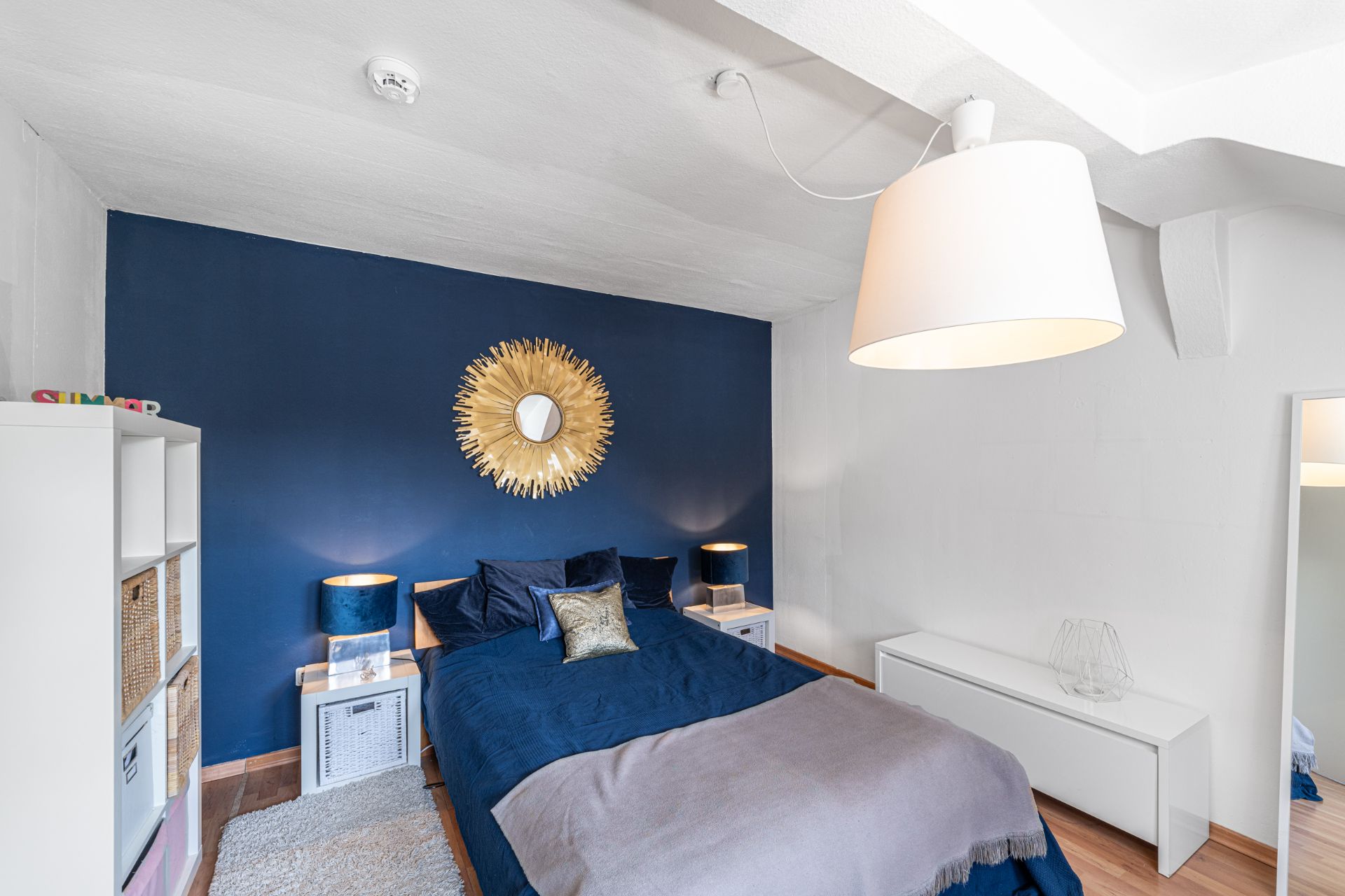 Modernes Schlafzimmer mit blauem Bett, goldener Spiegel an blauer Wand und großer weißer Lampe.
