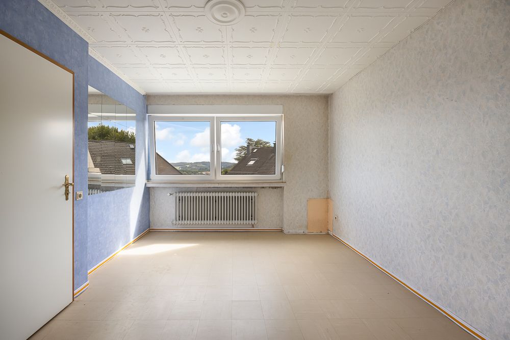Leeres Zimmer mit blau-grauen Wänden, großem Fenster und Heizkörper unter dem Fenster.