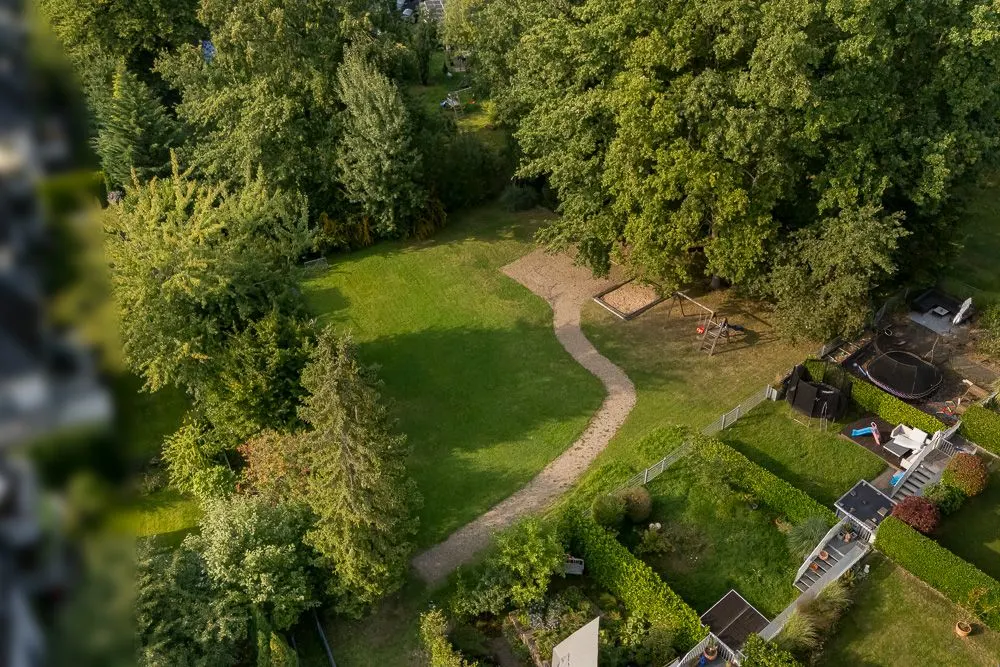 Großer Garten mit Rasen, Weg, Sandkasten und Kinderschaukel, umgeben von Bäumen.