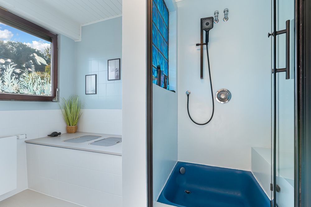 Modernes Bad mit Fenster, Glasdusche, blauer Duschwanne und Pflanze auf der Ablage.