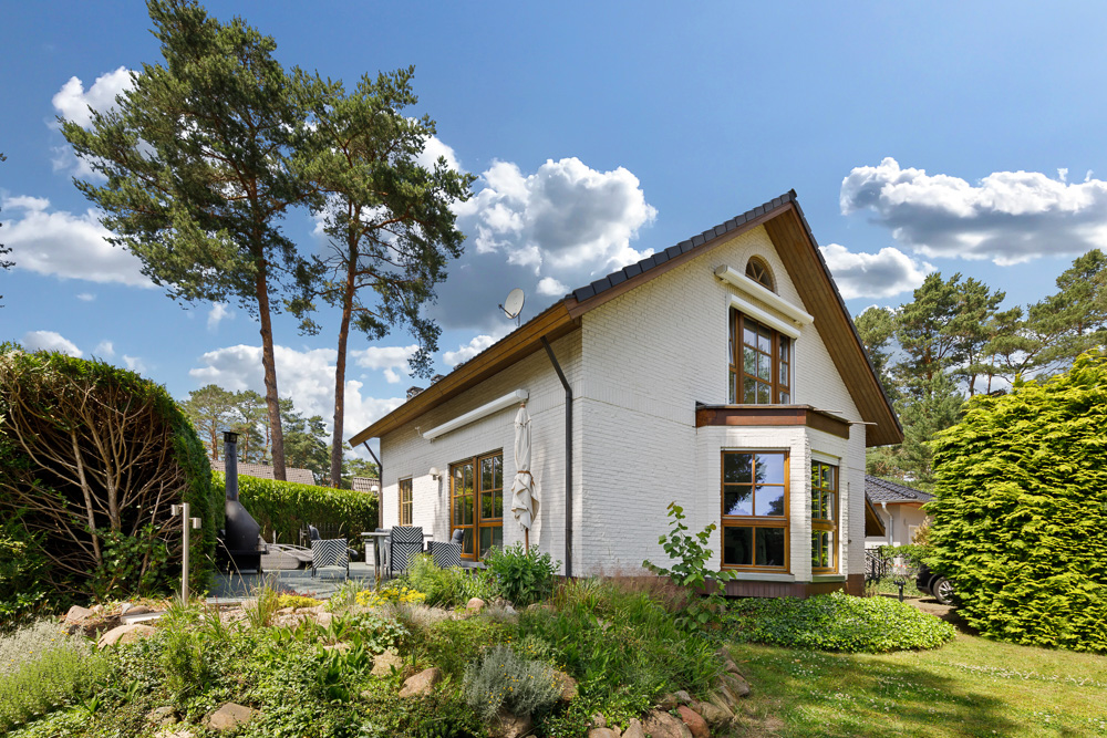 Weißes Einfamilienhaus mit Garten, Terrasse und großen Fenstern, umgeben von Bäumen und Hecken.
