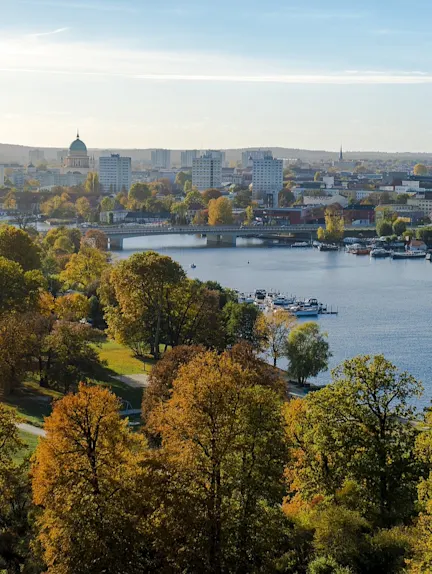 Blick auf eine Stadt mit Fluss, Brücke, vielen Bäumen und Gebäuden im Hintergrund.