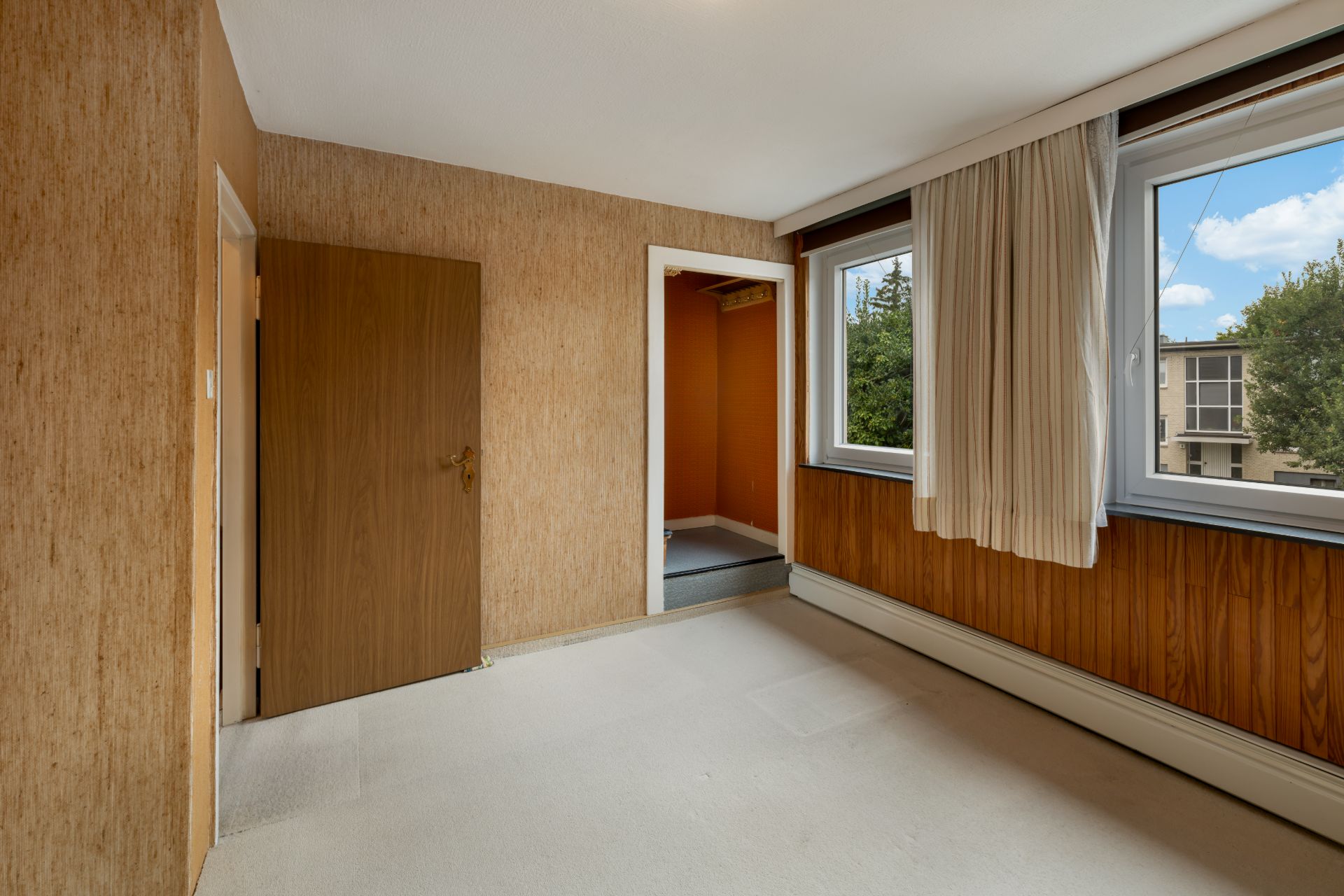 Leeres Zimmer mit braunen Wänden, offenem Türrahmen und zwei Fenstern mit Vorhängen.