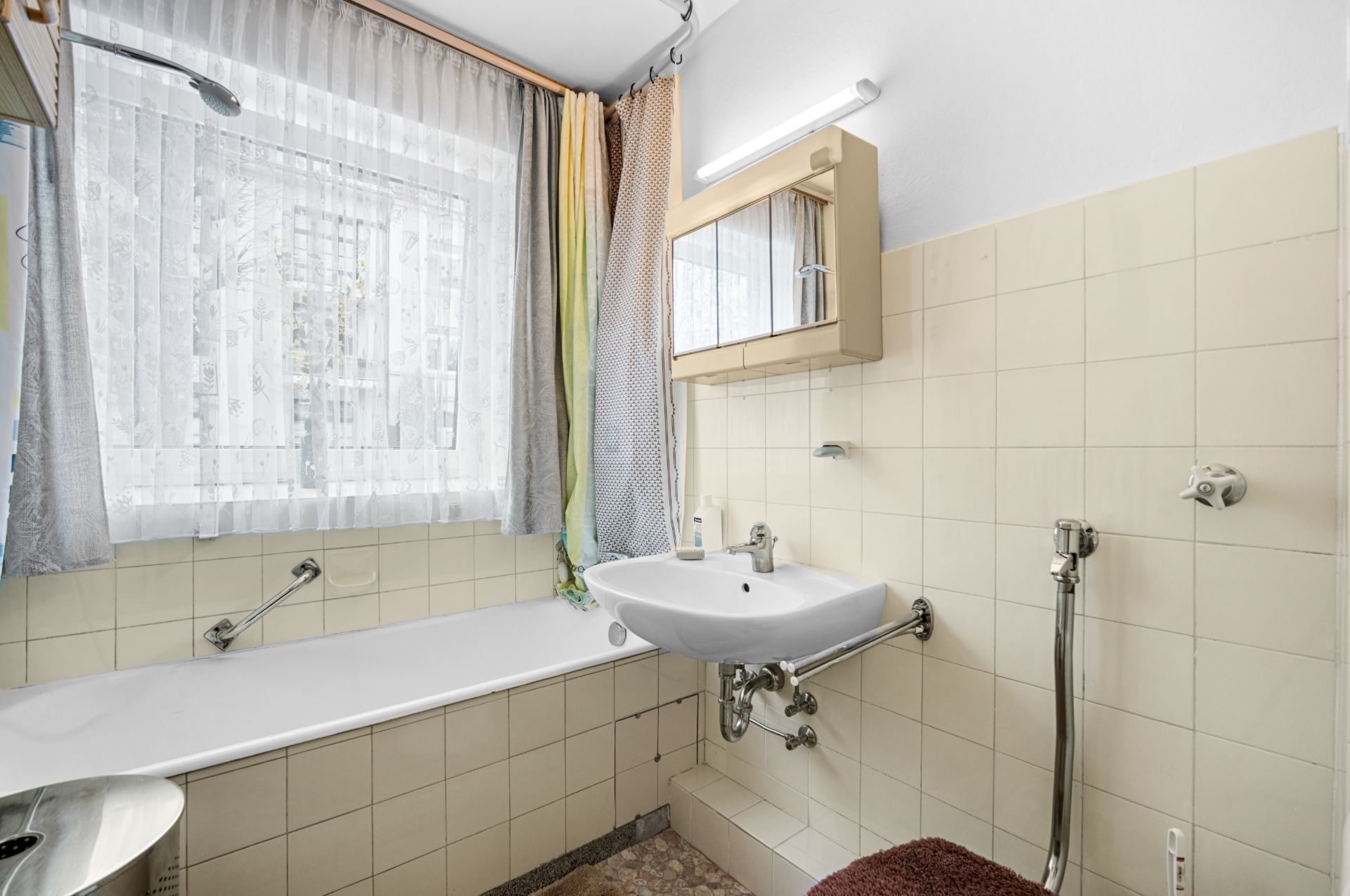 Helles Badezimmer mit Badewanne, Waschbecken, Spiegel und Fenster mit Vorhängen.