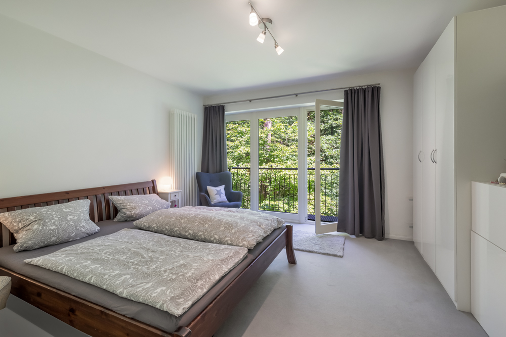 Helles Schlafzimmer mit Doppelbett, Sessel, weißem Schrank und offener Balkontür mit Blick ins Grüne.