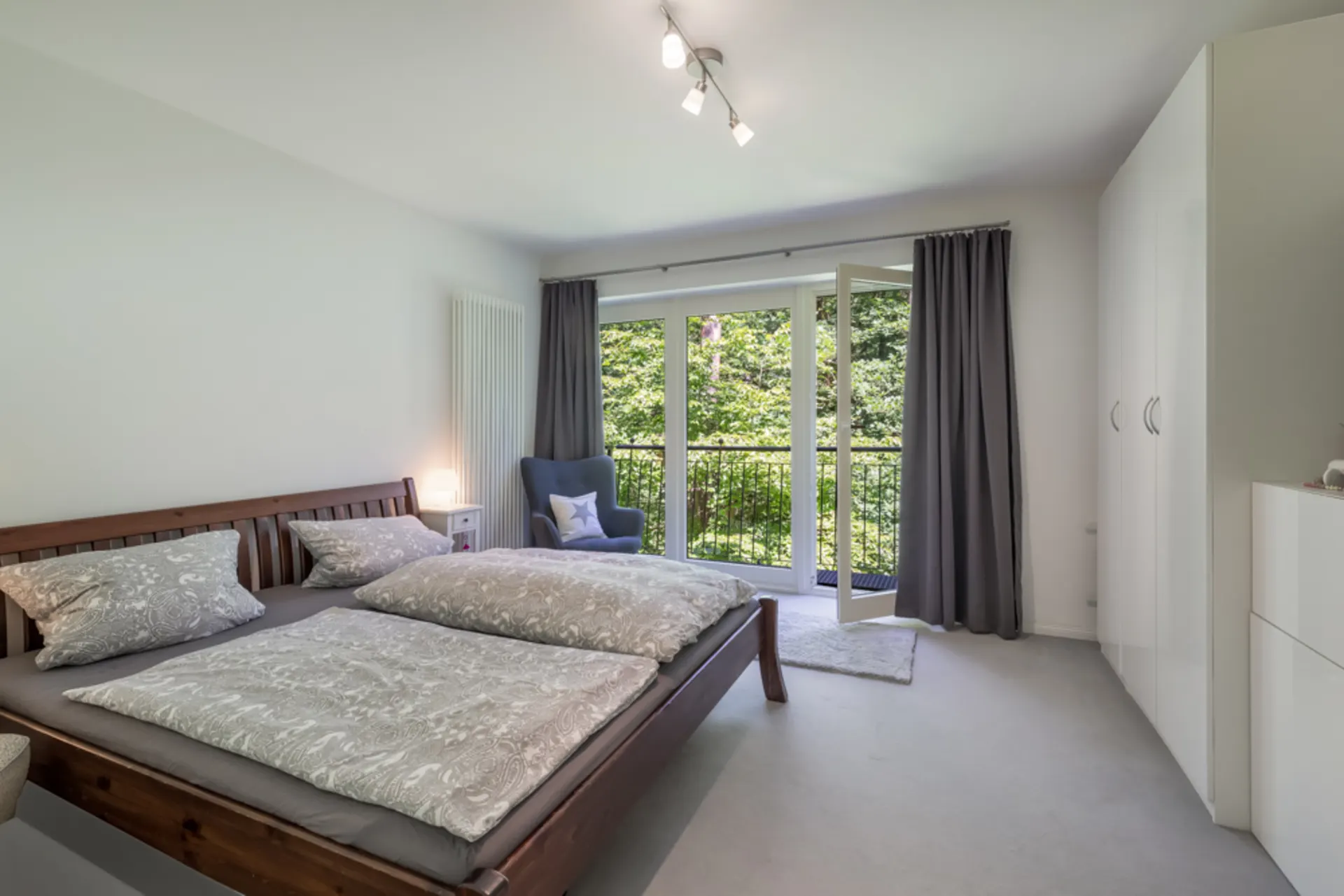 Helles Schlafzimmer mit Doppelbett, Sessel, weißem Schrank und offener Balkontür mit Blick ins Grüne.