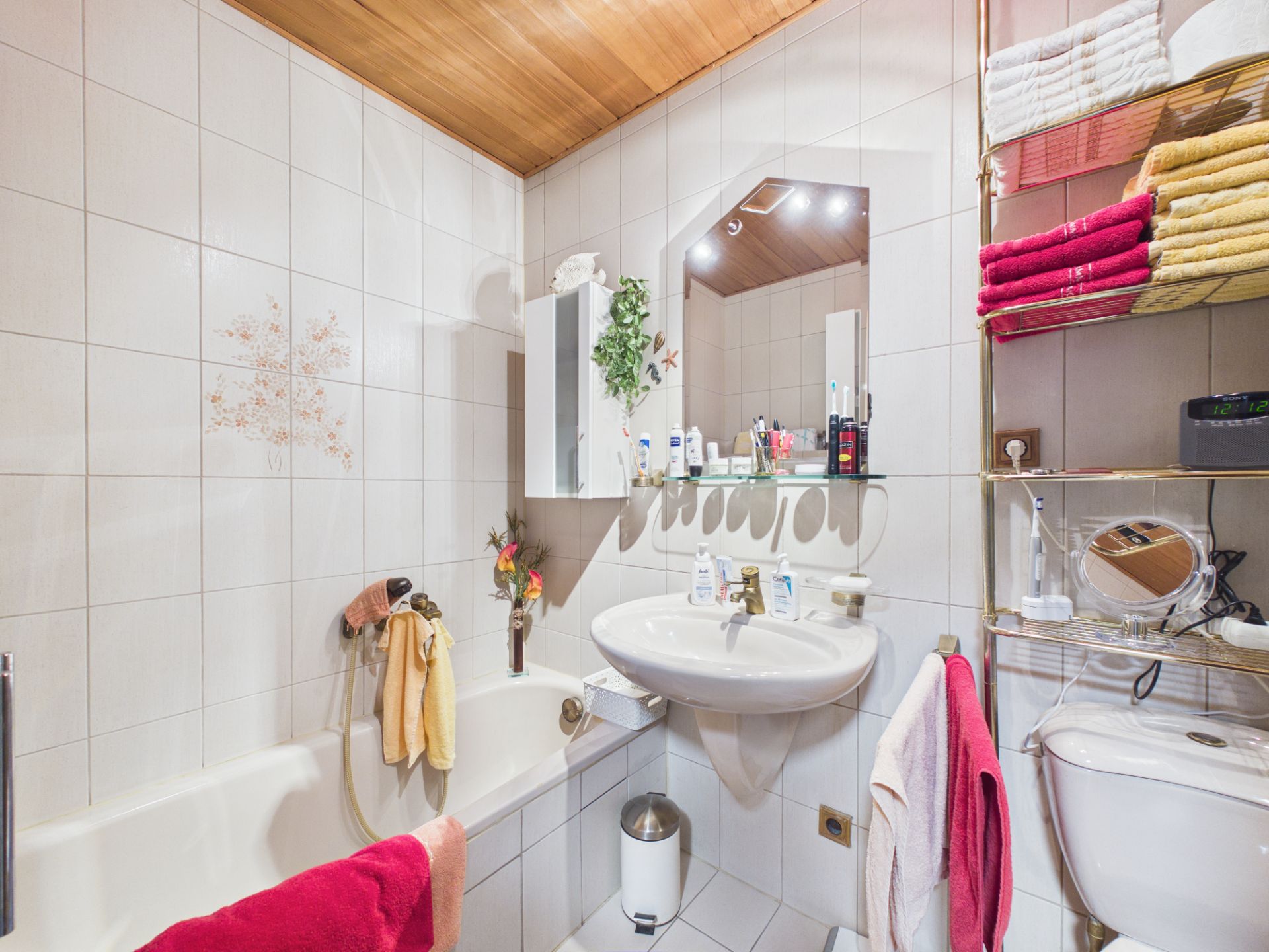 Kleines Badezimmer mit Badewanne, Waschbecken, Spiegel und Regal mit bunten Handtüchern.