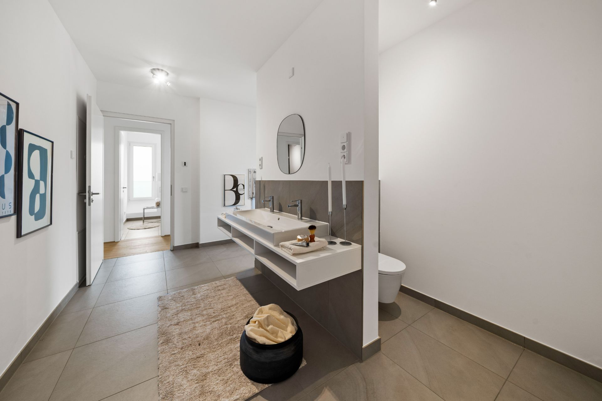 Modernes Badezimmer mit Doppelwaschbecken, rundem Spiegel und beigem Teppich auf Fliesenboden.