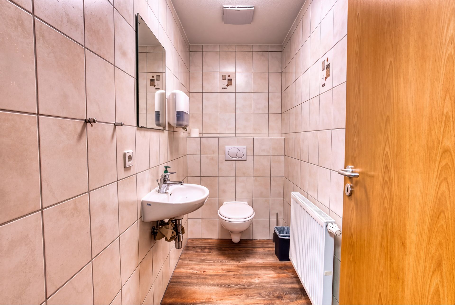 Gefliestes Badezimmer mit Waschbecken, Spiegel, Toilette, Heizkörper und Abfalleimer.
