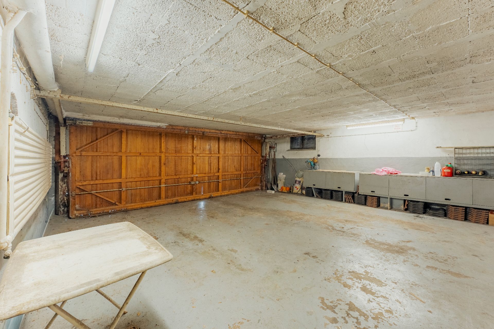 Leere Garage mit Holztor, Tisch links und Regalen mit verschiedenen Gegenständen an der rechten Wand.