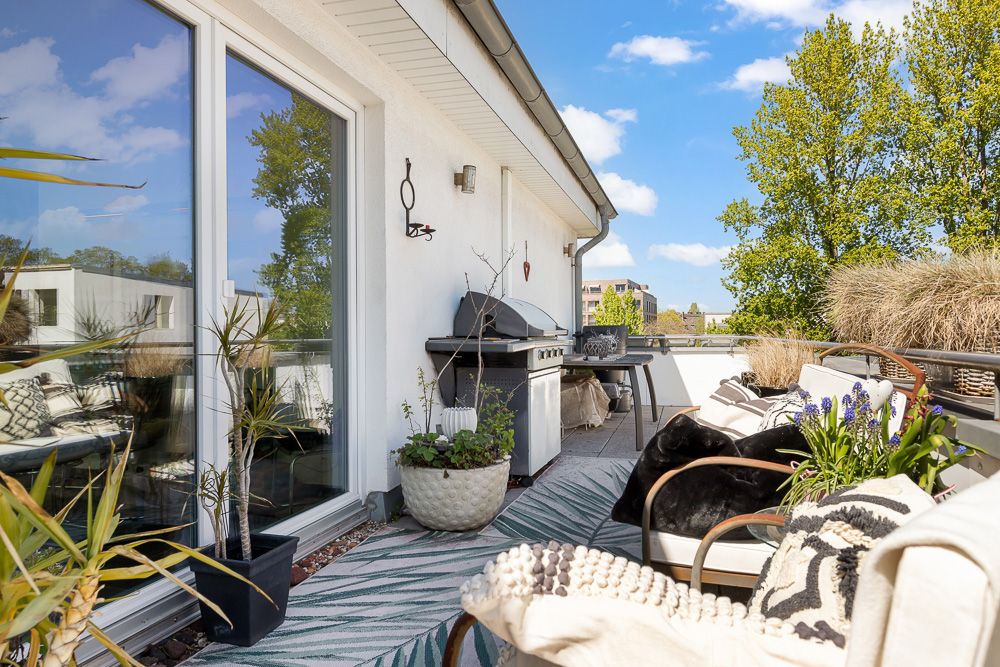 Möblierte Terrasse mit Grill, Pflanzen, Sitzgelegenheiten und Blick auf Bäume bei sonnigem Wetter.