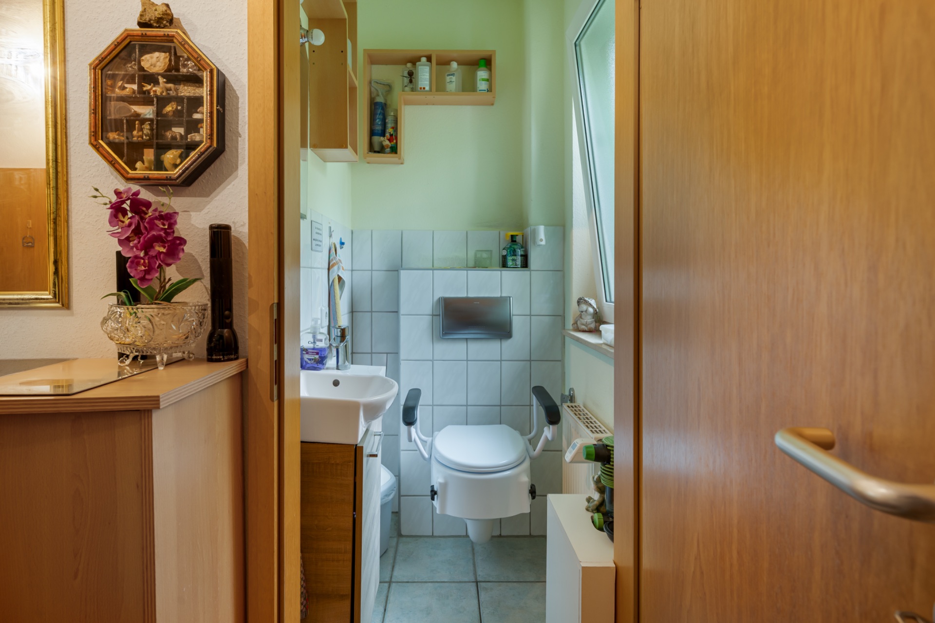 Kleines Badezimmer mit Toilette, Haltegriffen, Waschbecken und offenem Fenster.