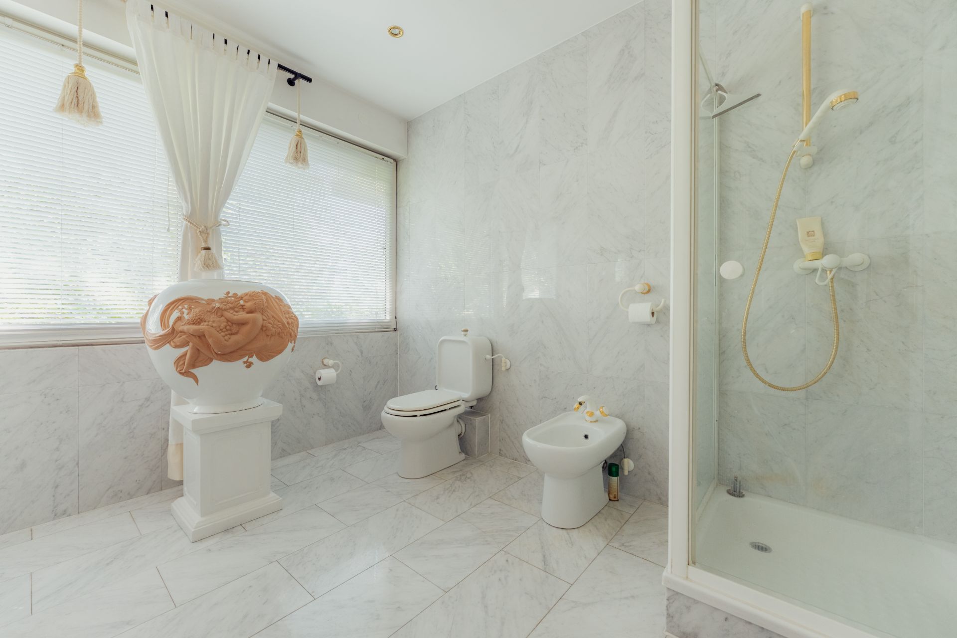 Helles Badezimmer mit Dusche, Toilette, Bidet und großer Vase vor Fenster.
