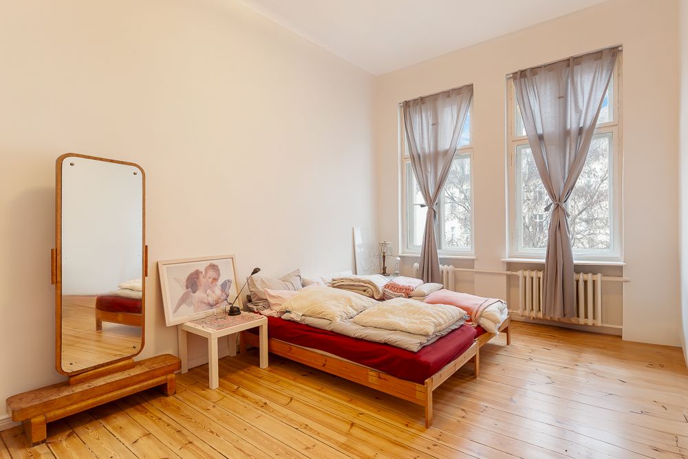 Schlafzimmer mit Holzboden, einfachem Bett, großem Spiegel und zwei Fenstern mit grauen Vorhängen.