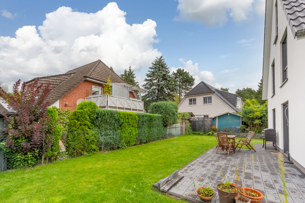 Garten mit Terrasse, Holzstühlen und Pflanzen neben einem Wohnhaus bei sonnigem Wetter.