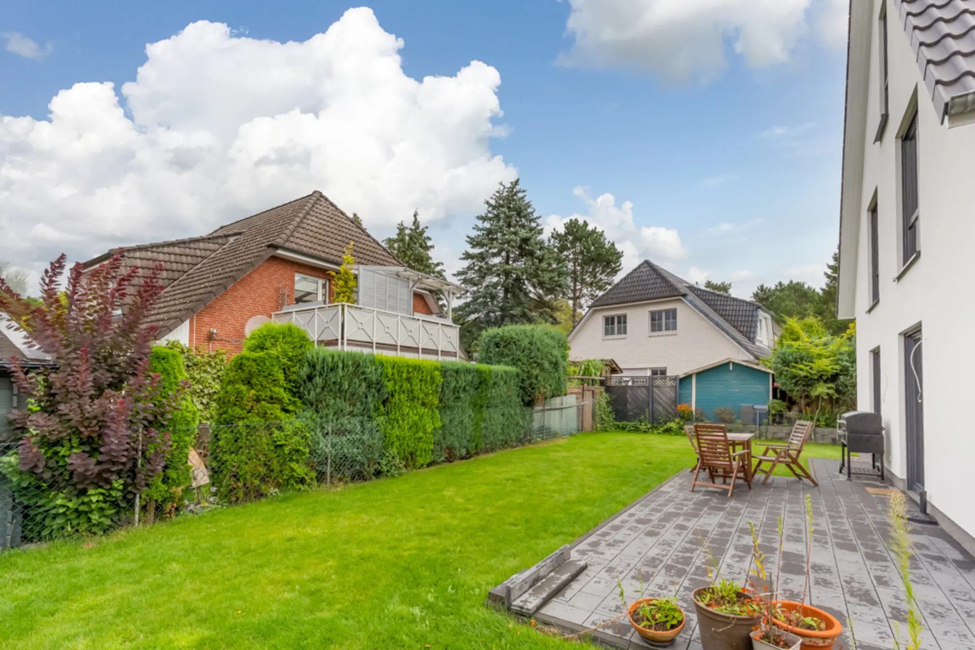 Garten mit Terrasse, Holzstühlen und Pflanzen neben einem Wohnhaus bei sonnigem Wetter.