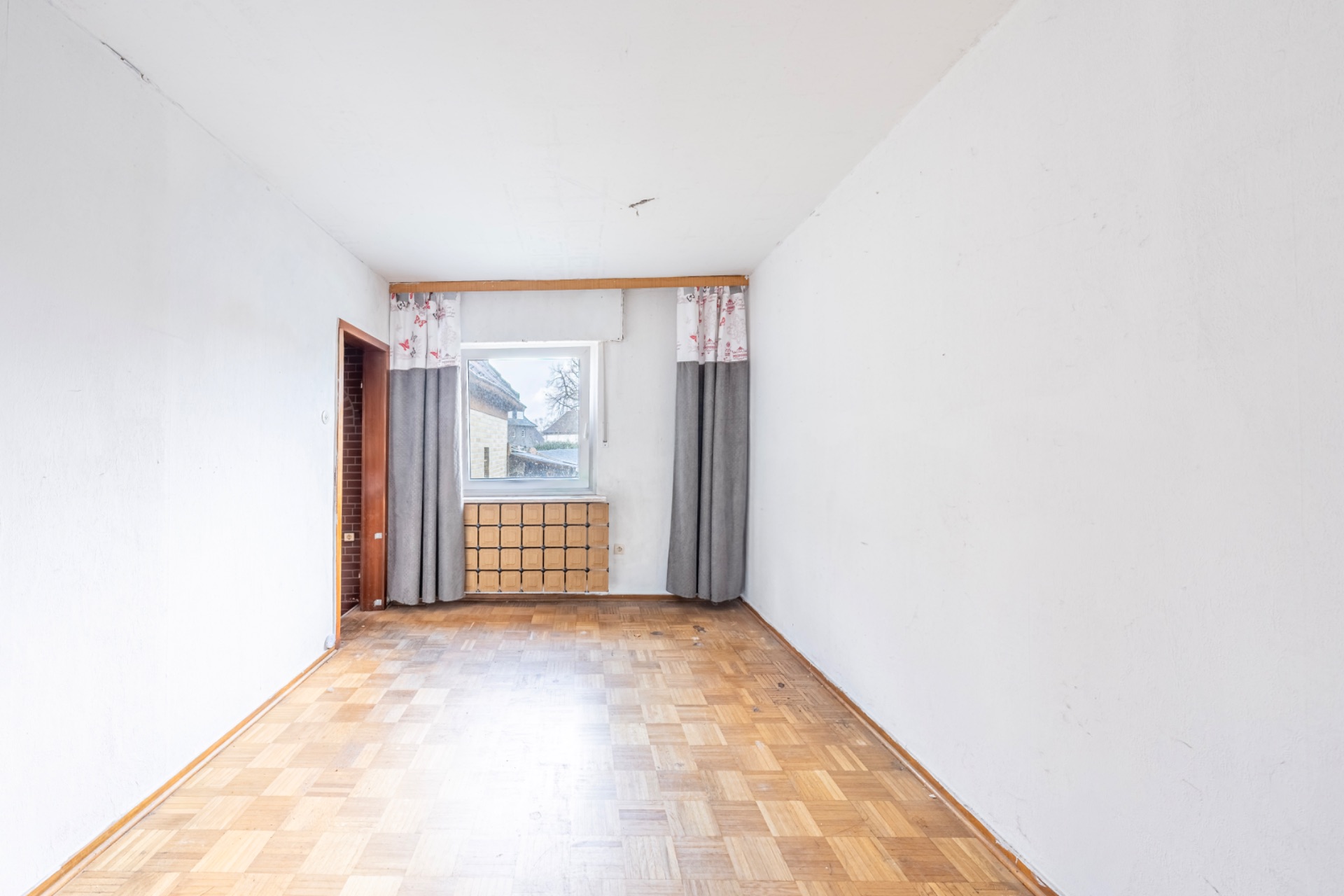 Leerer Raum mit Parkettboden, Fenster mit Vorhängen und Heizung unter dem Fenster.