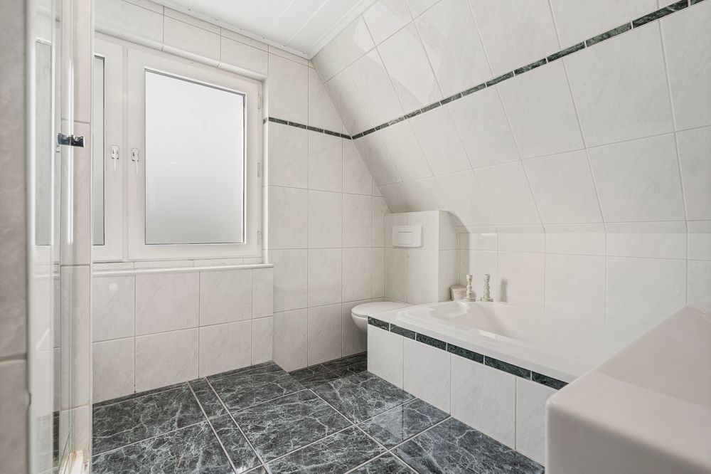 Helles Badezimmer mit Badewanne, Toilette und großem Fenster, Fliesen an Wand und Boden.