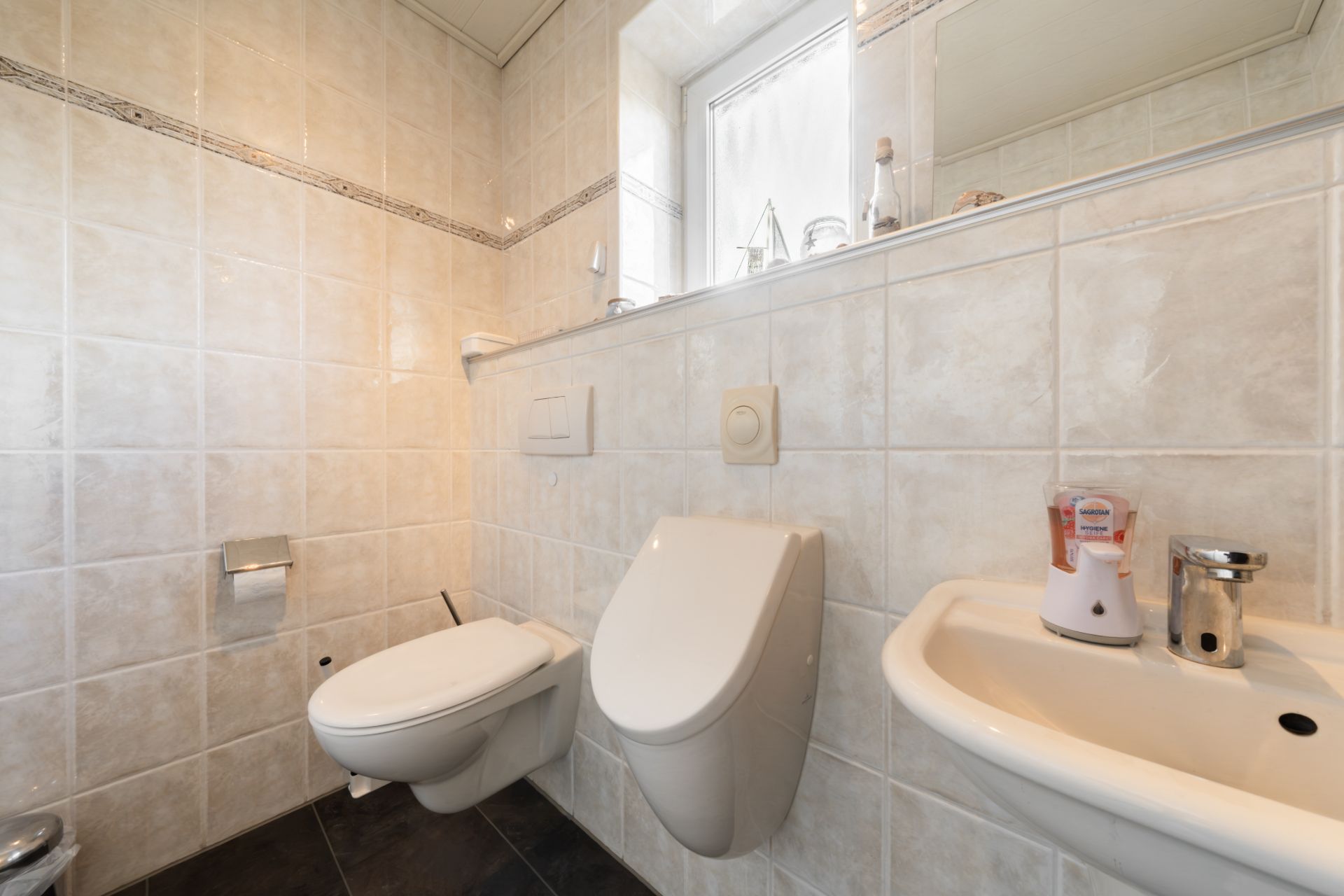 Helles Badezimmer mit WC, Urinal und kleinem Waschbecken vor gefliester Wand und Fenster.
