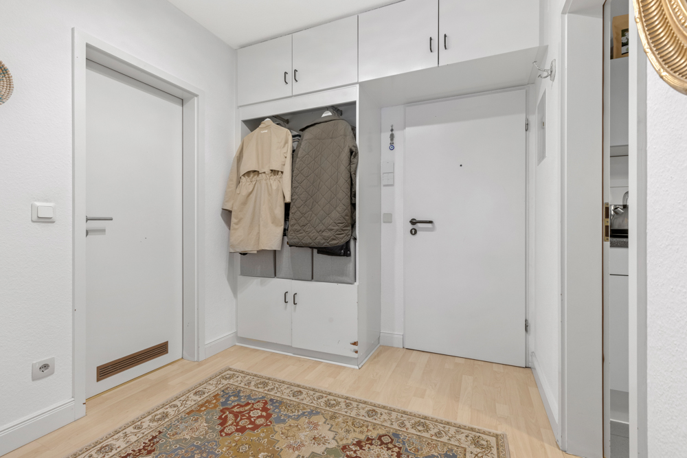 Heller Flur mit Garderobe, aufgehängten Jacken, weißen Türen und Teppich auf Holzboden.