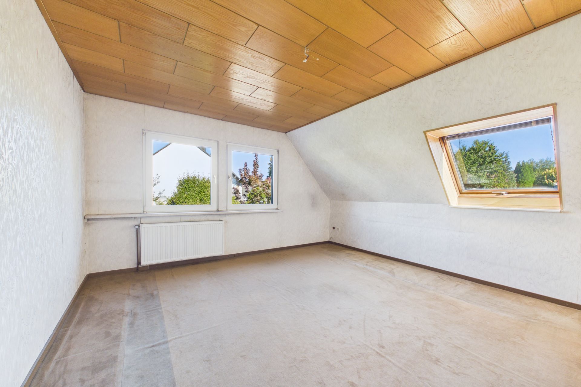 Leeres Zimmer mit schräger Decke, Teppichboden und zwei Fenstern, eines davon ist ein Dachfenster.