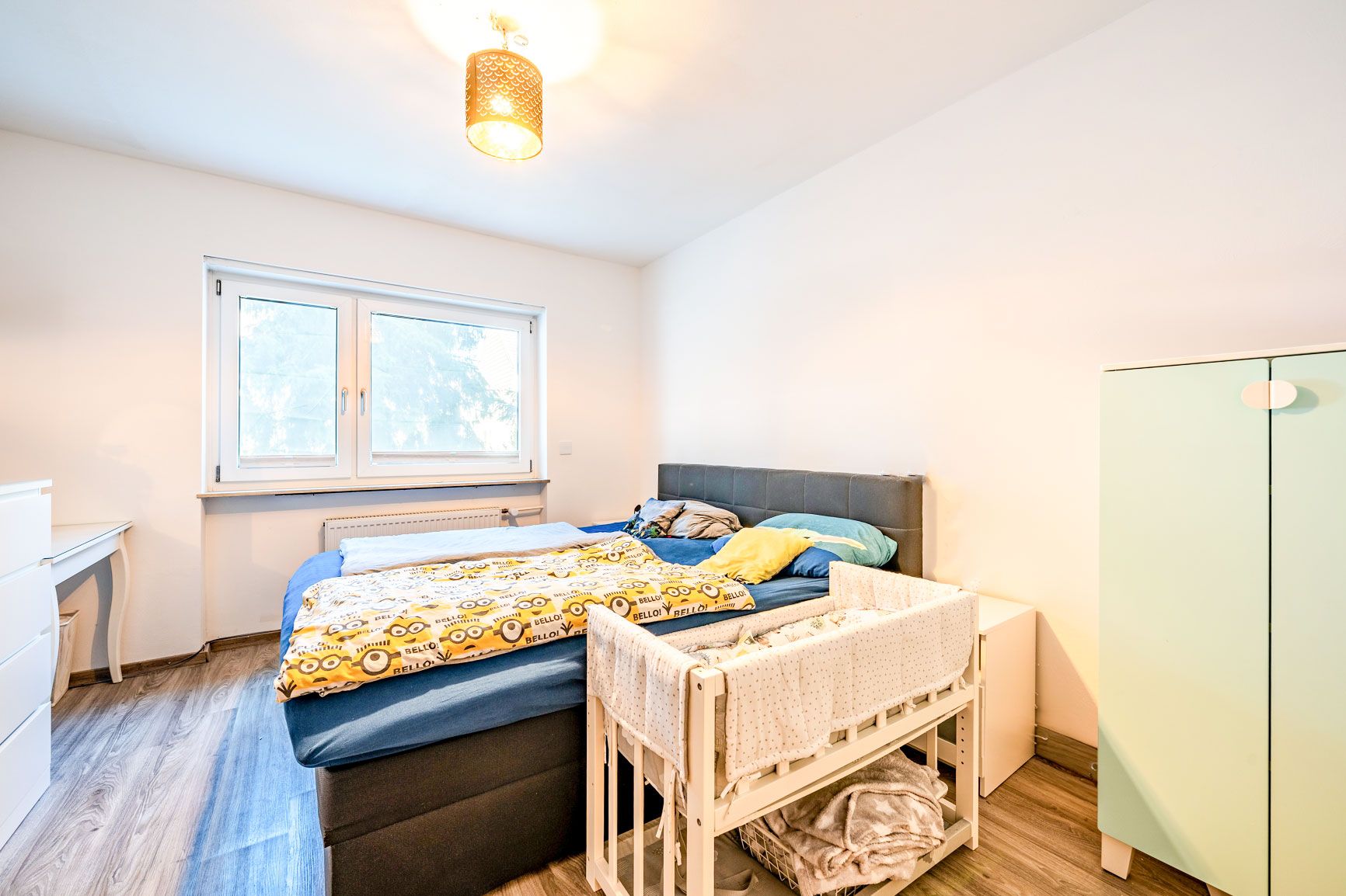 Helles Schlafzimmer mit Doppelbett, Babybett, Schrank und Fenster.