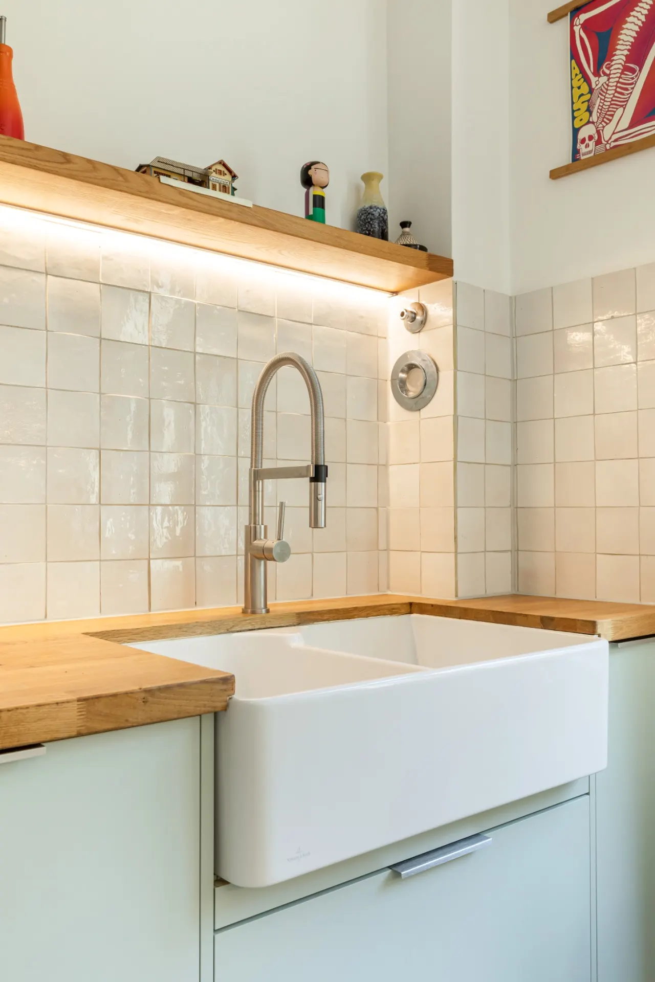 Helle Küche mit weißer Keramikspüle, Holzboden und Fliesenspiegel, Regal mit Dekorationen darüber.