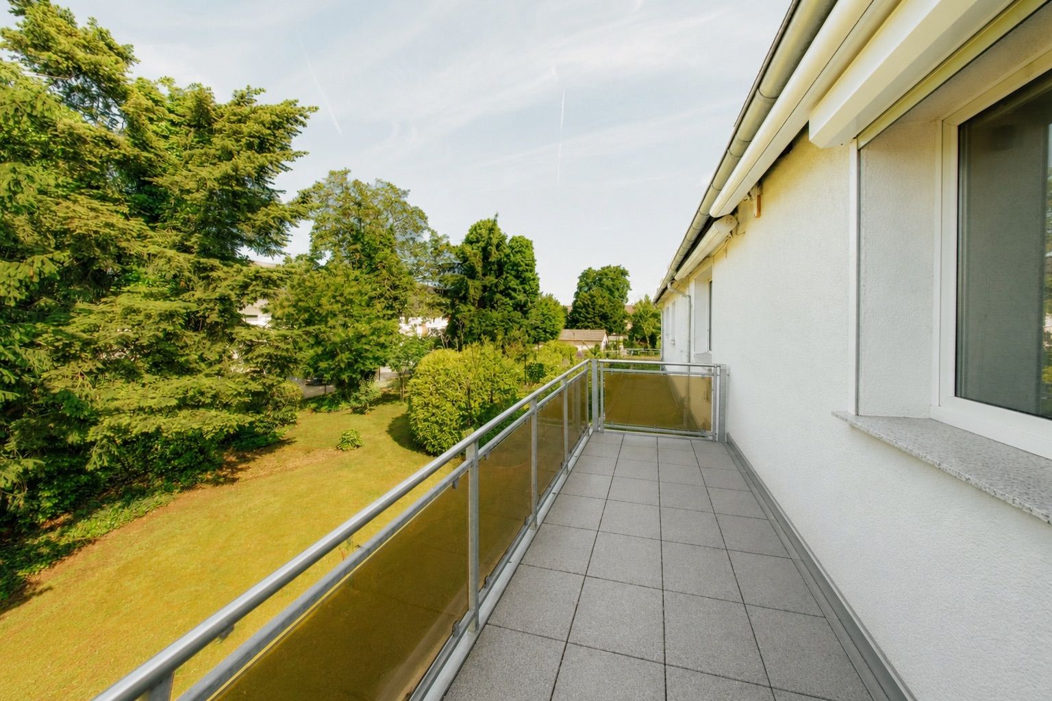 Langer Balkon mit Fliesenboden, Glasgeländer und Blick auf einen grünen Garten mit Bäumen.