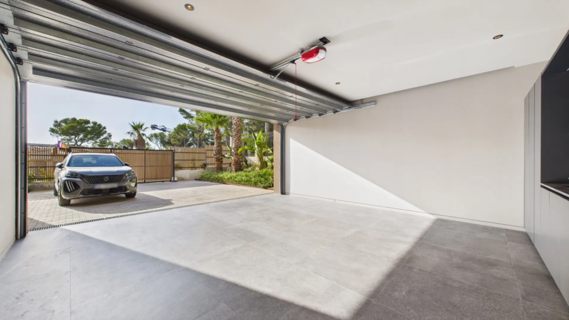 Leere Garage mit offenem Tor, draußen steht ein Auto und es sind Bäume im Hintergrund sichtbar.