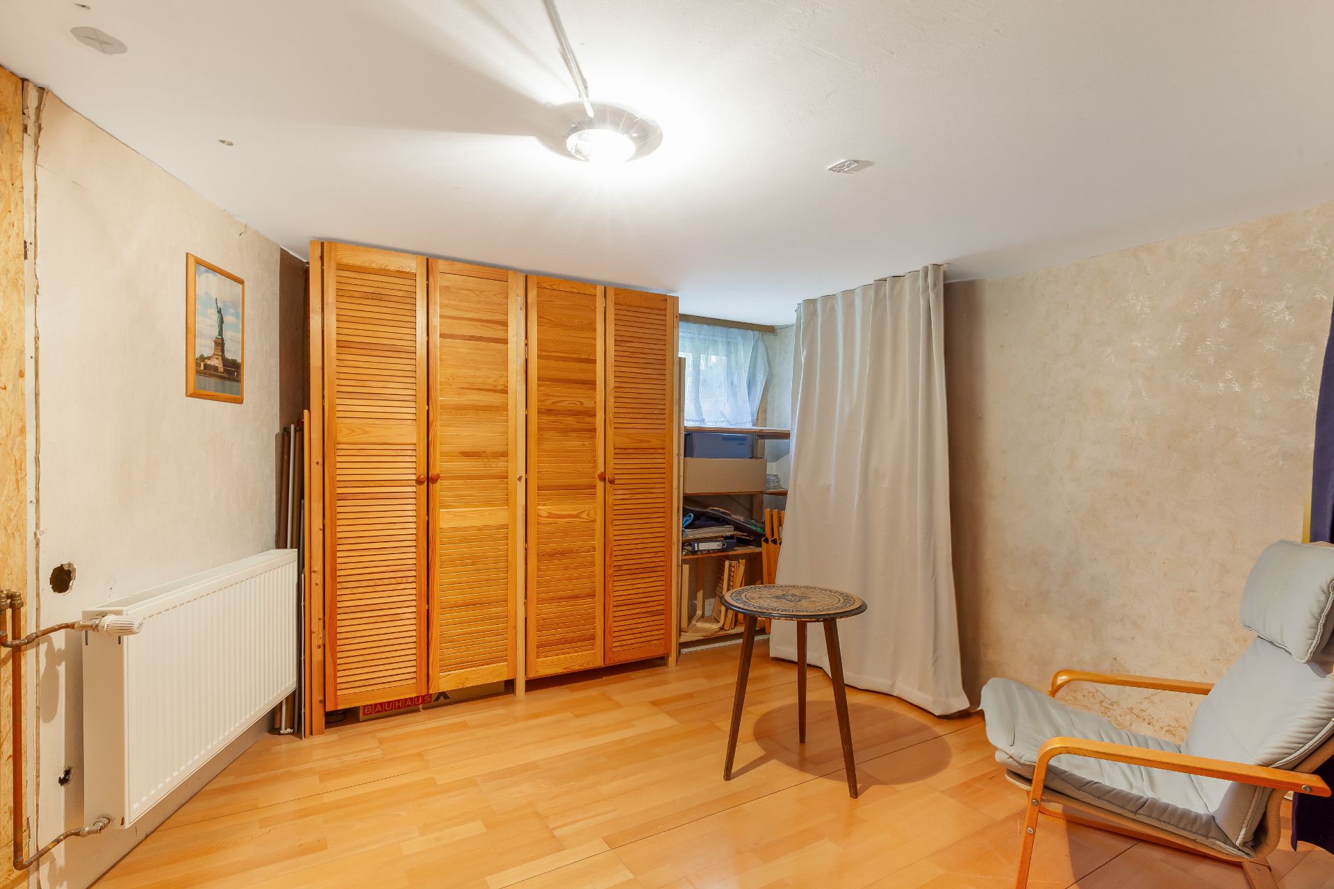 Zimmer mit Holzschrank, Sessel, kleinem Tisch und Fenster mit Vorhang.