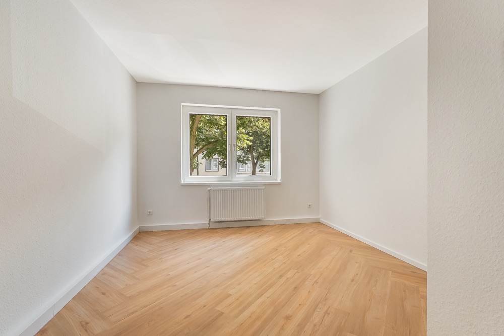 Helles, leeres Zimmer mit Holzfußboden und großem Fenster, durch das Bäume zu sehen sind.