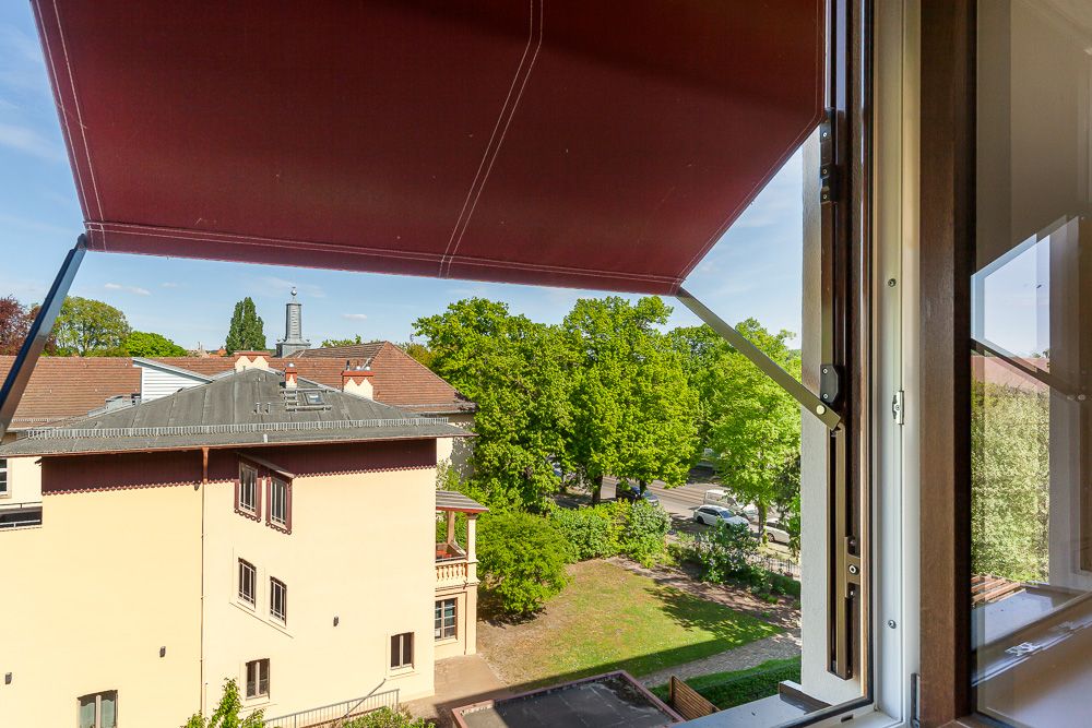Blick aus einem Fenster mit roter Markise auf Nachbarhaus und grüne Bäume.
