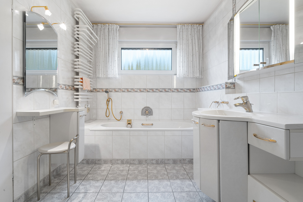 Helles Badezimmer mit Badewanne, zwei Fenstern, Waschbecken und Handtuchheizkörper.